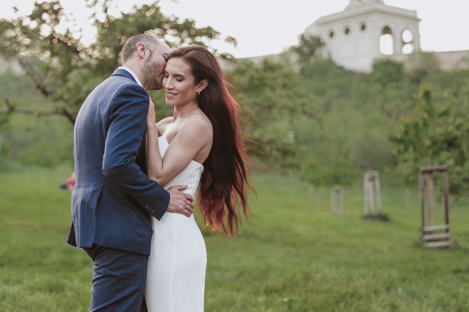 Bride in a white dress and groom in a navy suit embrace in a green meadow with trees and a hilltop building in the background.