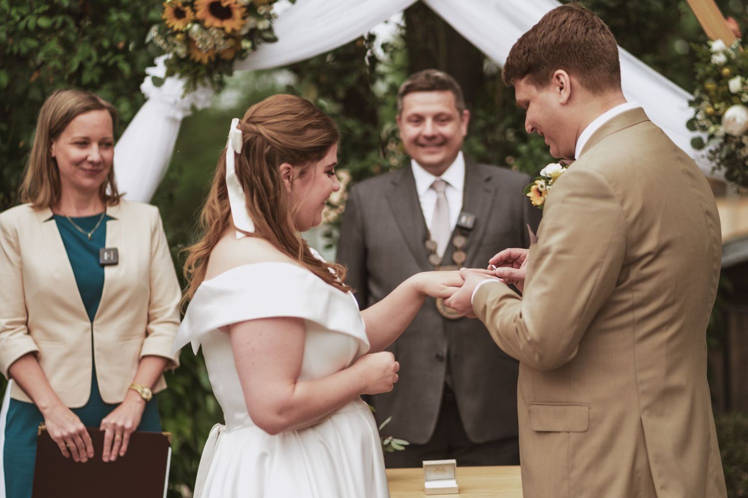 Bride in a white off-the-shoulder gown and groom in a tan suit exchange rings during an outdoor wedding ceremony as the officiant smiles.