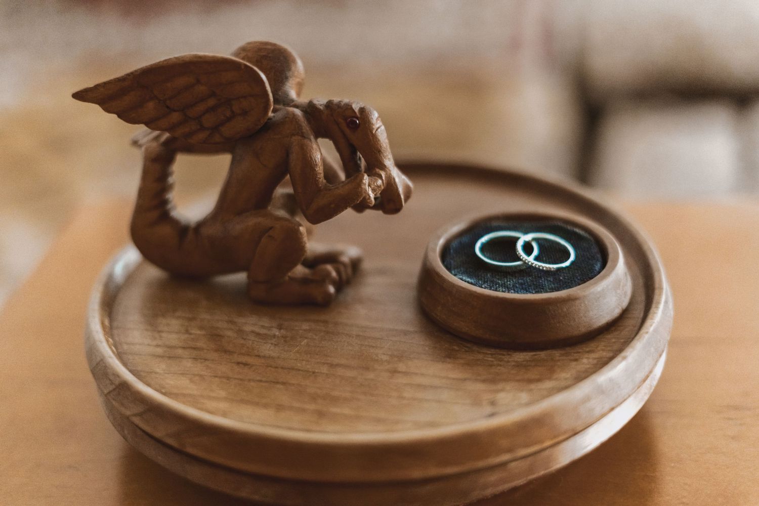 Carved wooden winged cherub figurine kneeling on a round wooden tray beside a small dark ring box holding two silver wedding bands.