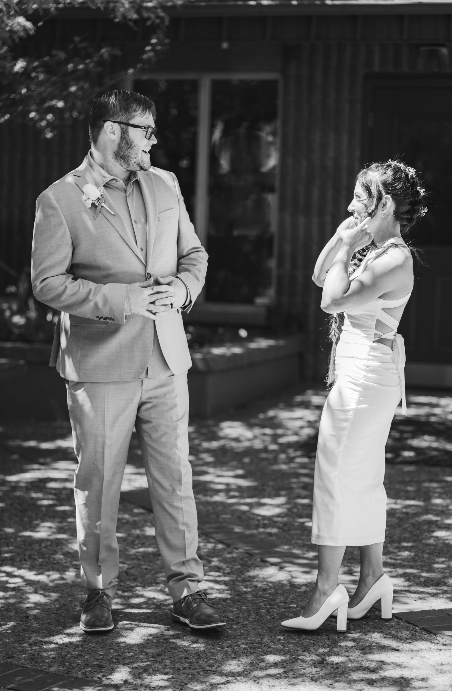 Black and white photo of a smiling bride in a white dress and heels sharing an emotional first look with a groom in a suit outdoors.