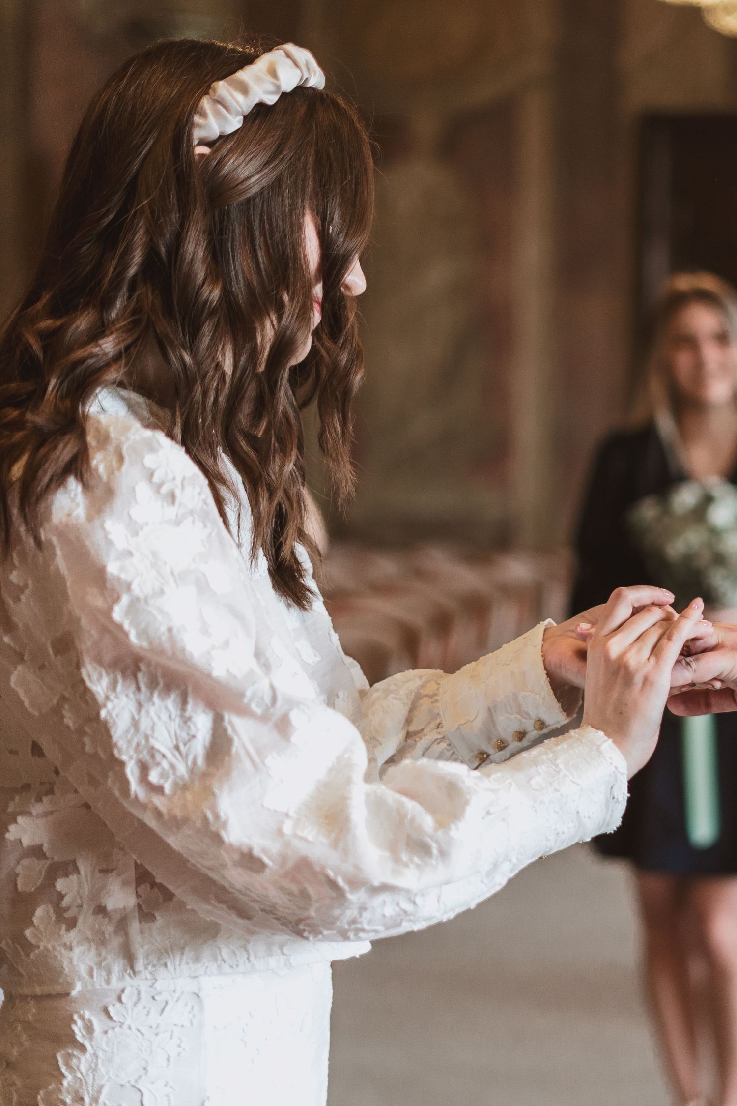Bride in a white lace dress and ruched headband exchanges rings during a wedding ceremony as a bridesmaid watches.