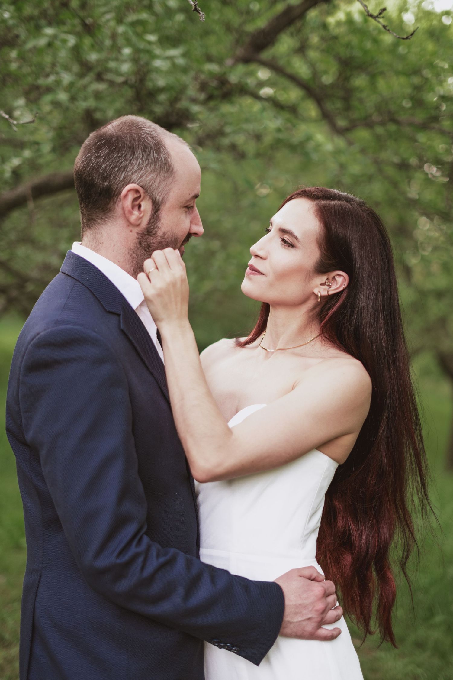Couple embracing in a green orchard, the man in a navy suit gently touching the face of a woman in a white dress with long auburn hair.