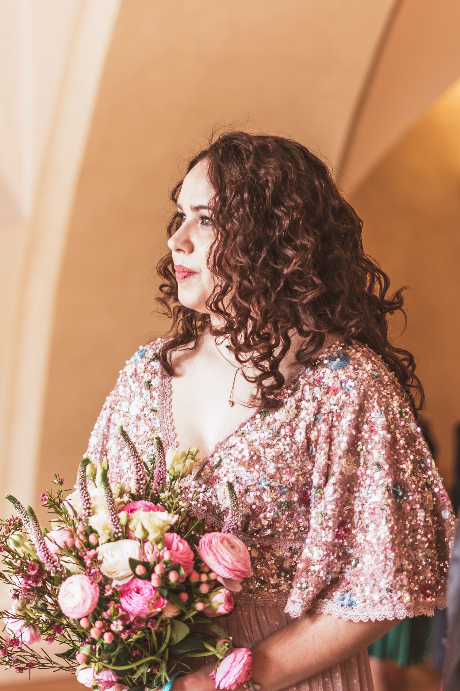 Bride with long curly brown hair in a pink sequin beaded gown holding a bouquet of pink and white roses, gazing to the side.