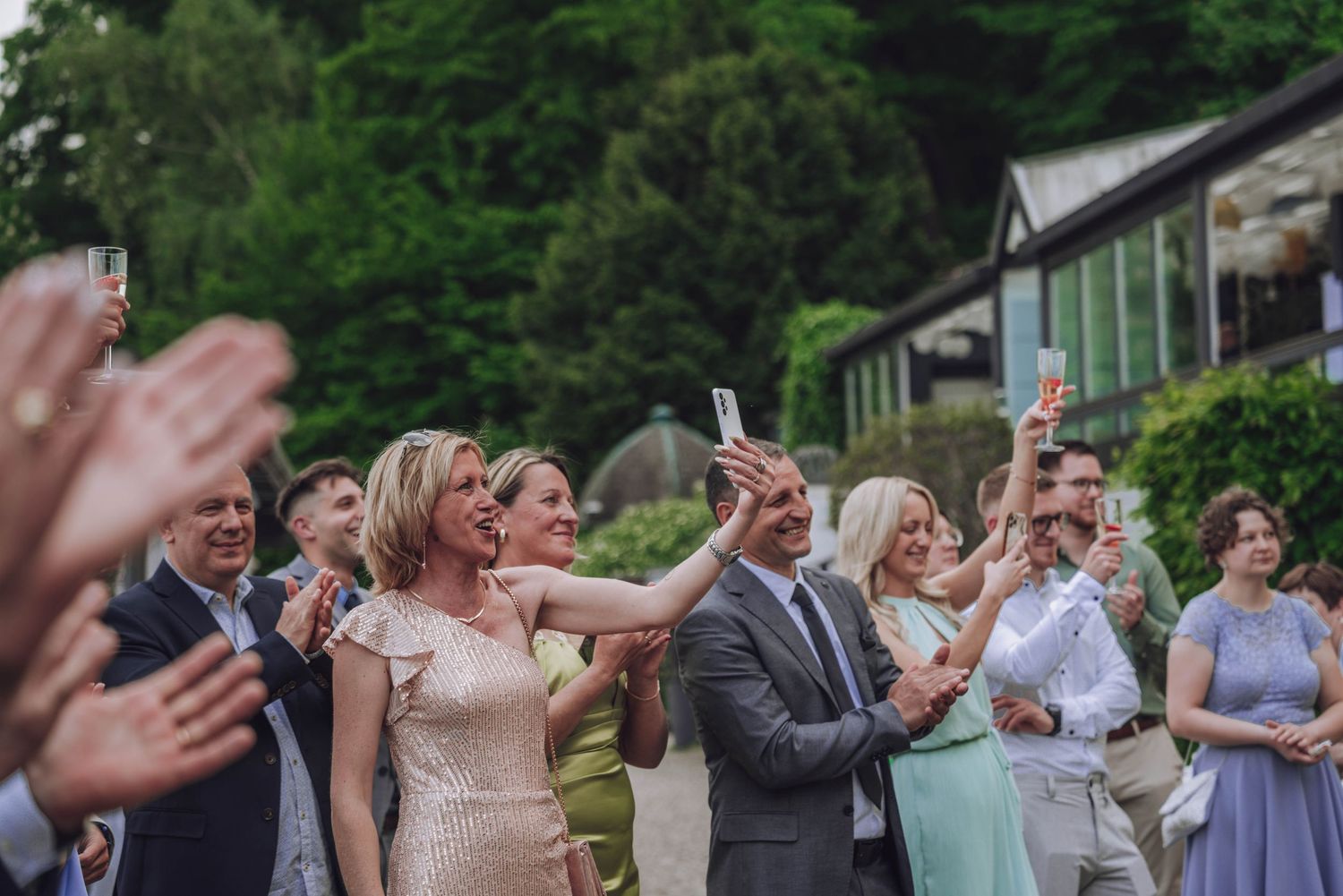 Wedding guests in formal attire cheer and clap outdoors, with one woman in a champagne dress raising her arm beside a leafy garden venue.