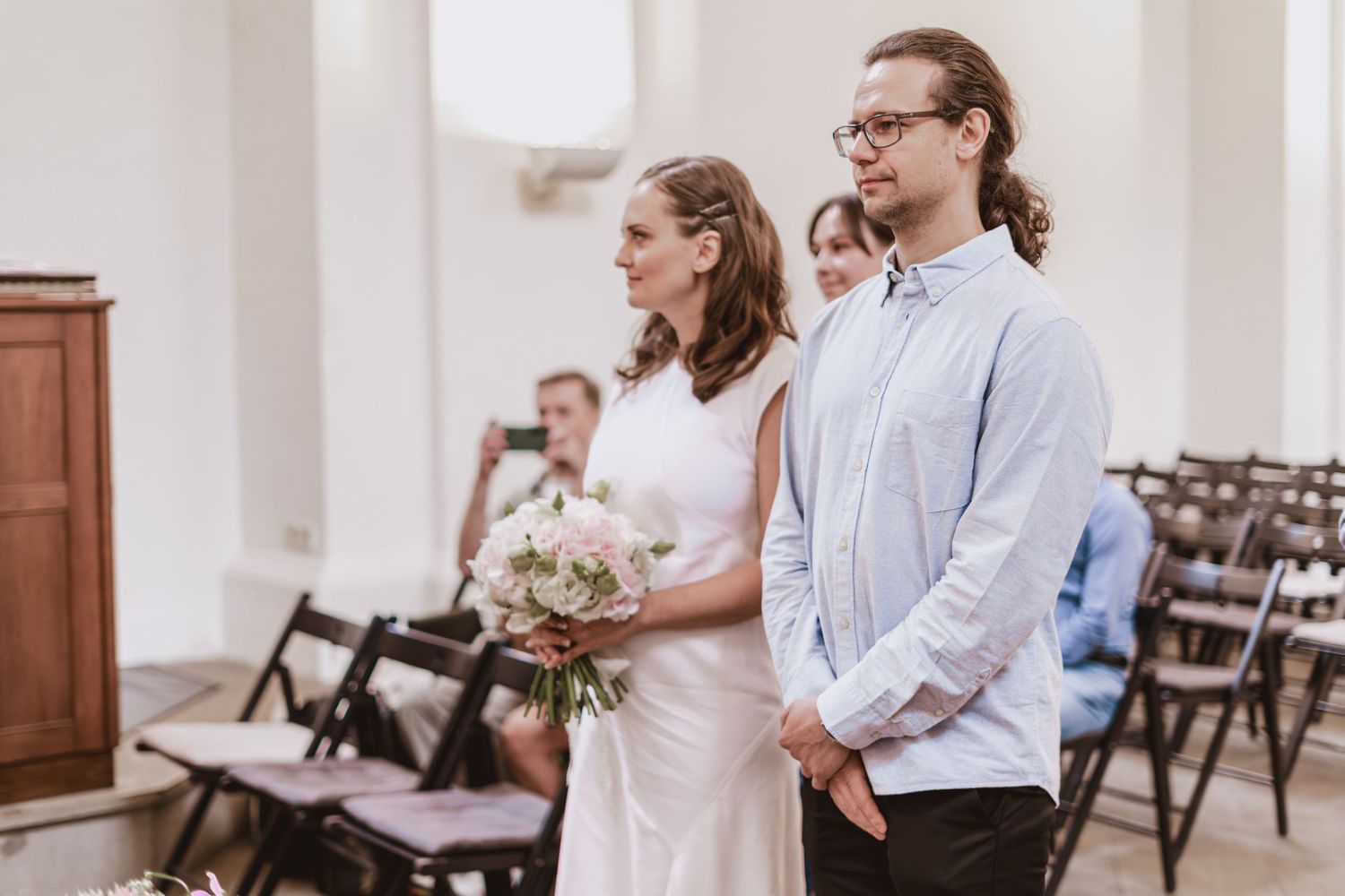 Bride in a white dress holding a bouquet stands beside a groom in a light blue shirt during a wedding ceremony with guests seated behind.