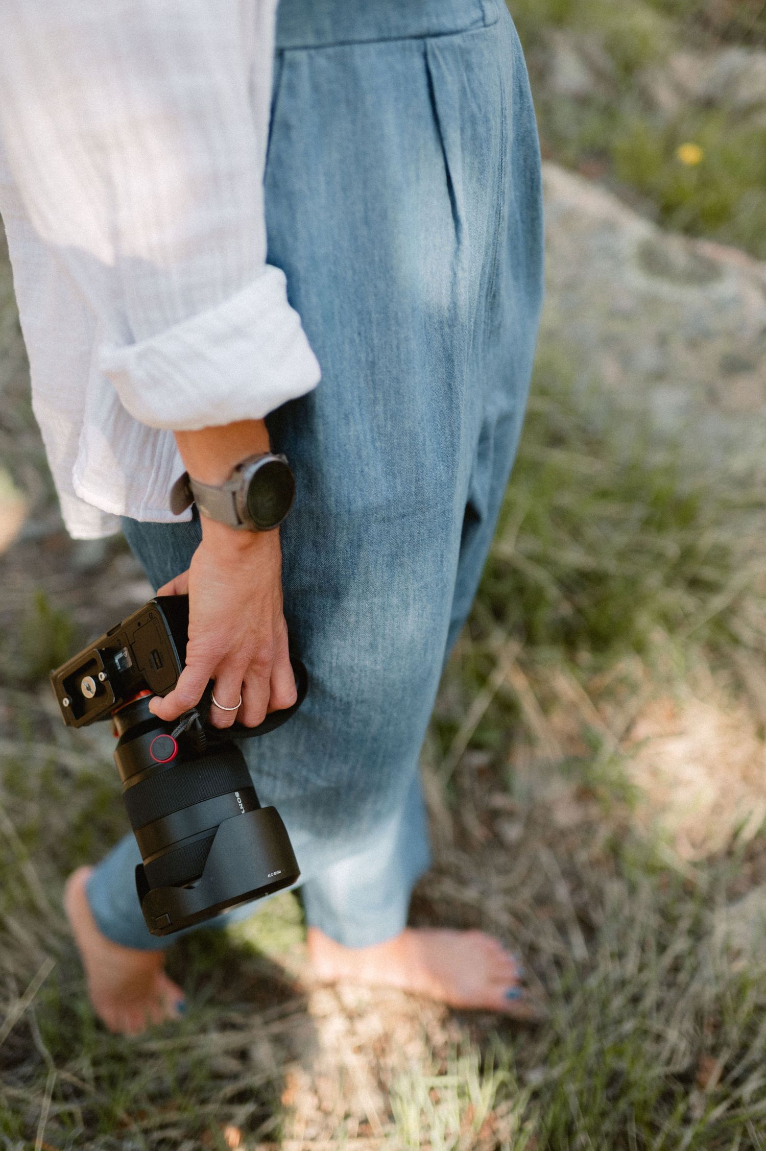 A hand holds a professional camera while wearing a flowy white top and light denim in an outdoor setting.