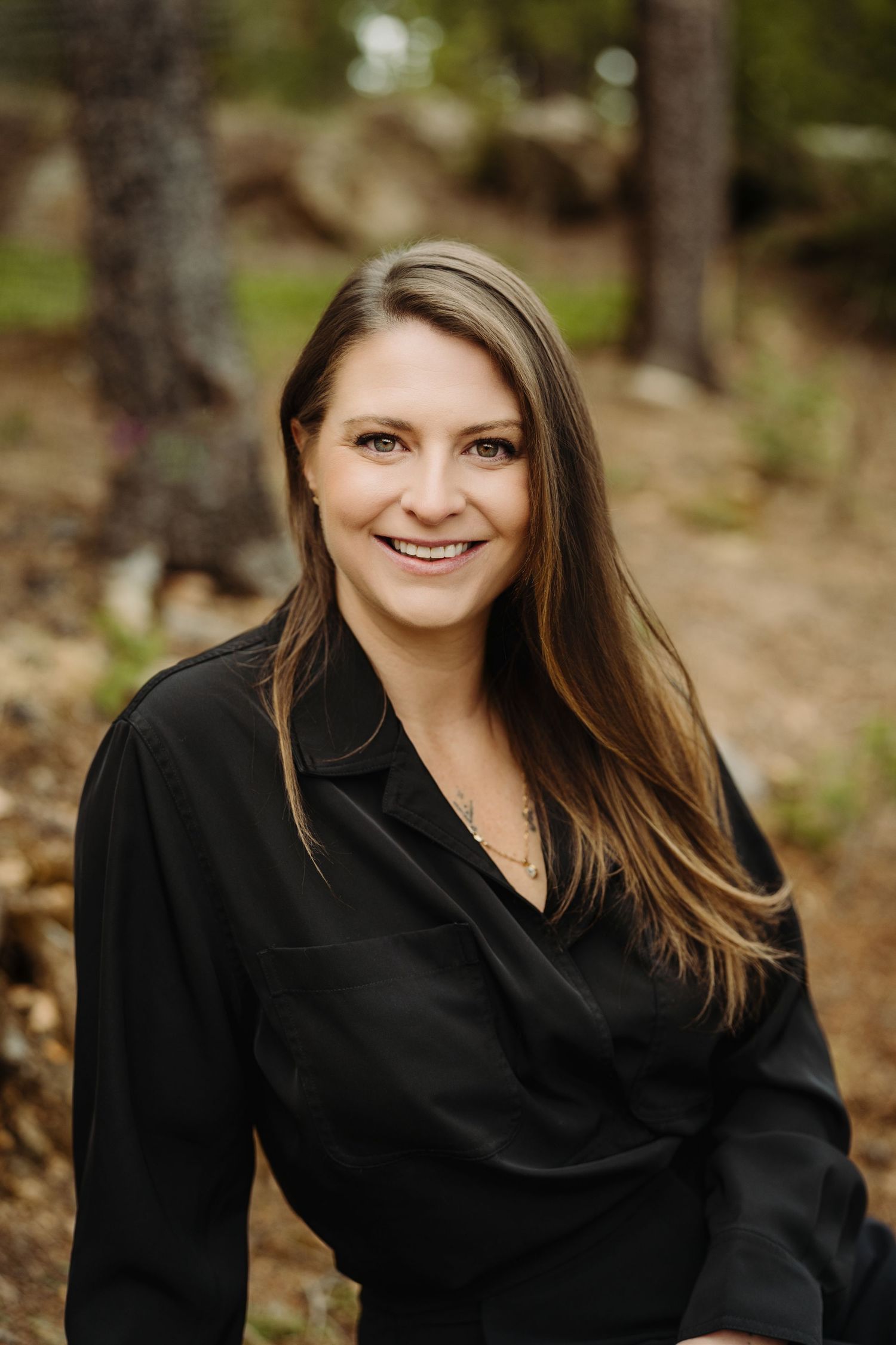 Smiling woman with long brown hair wearing a black blouse, posing outdoors in a forest setting.