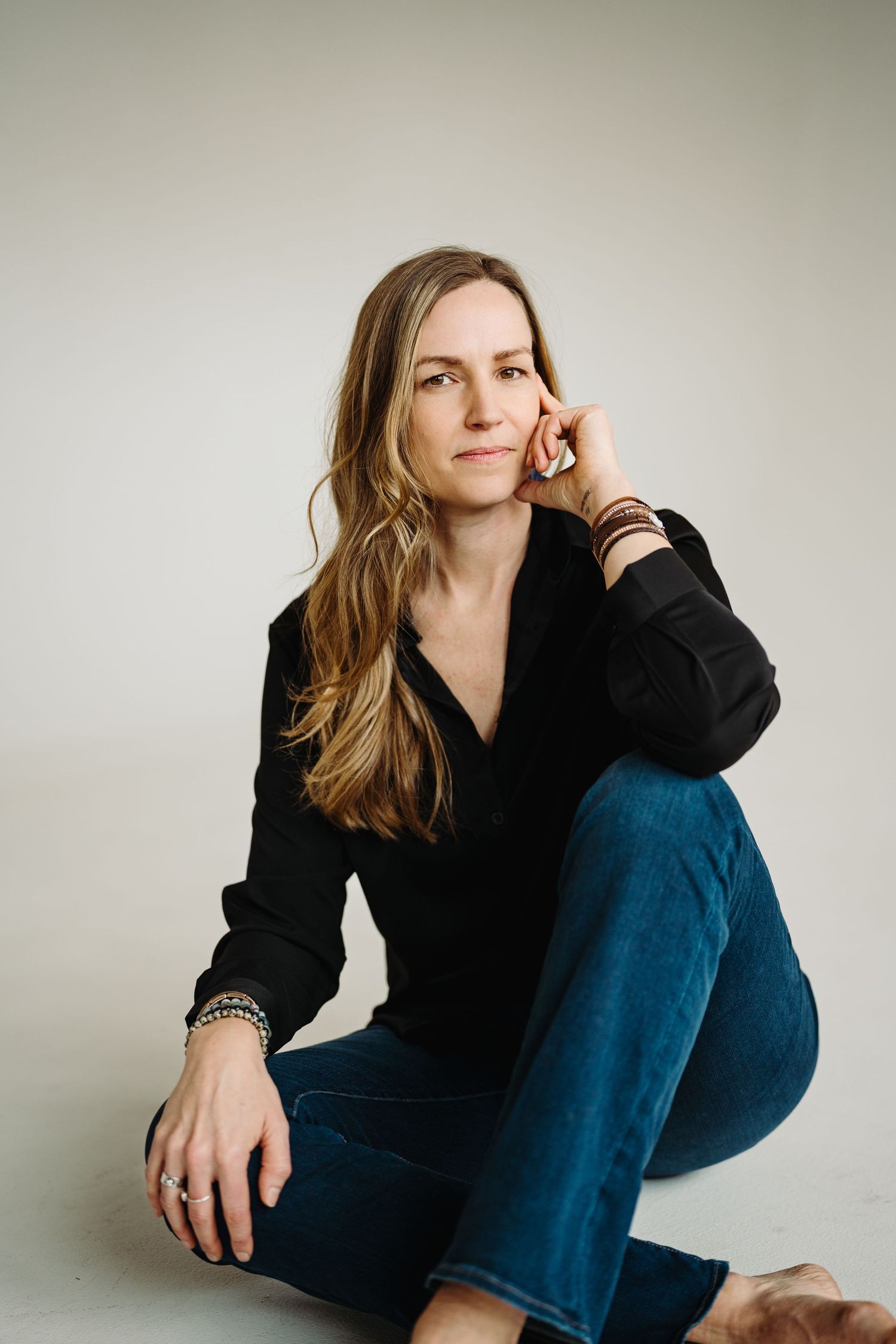 A casual portrait against white background wearing a black sweater and blue jeans in a relaxed seated pose.