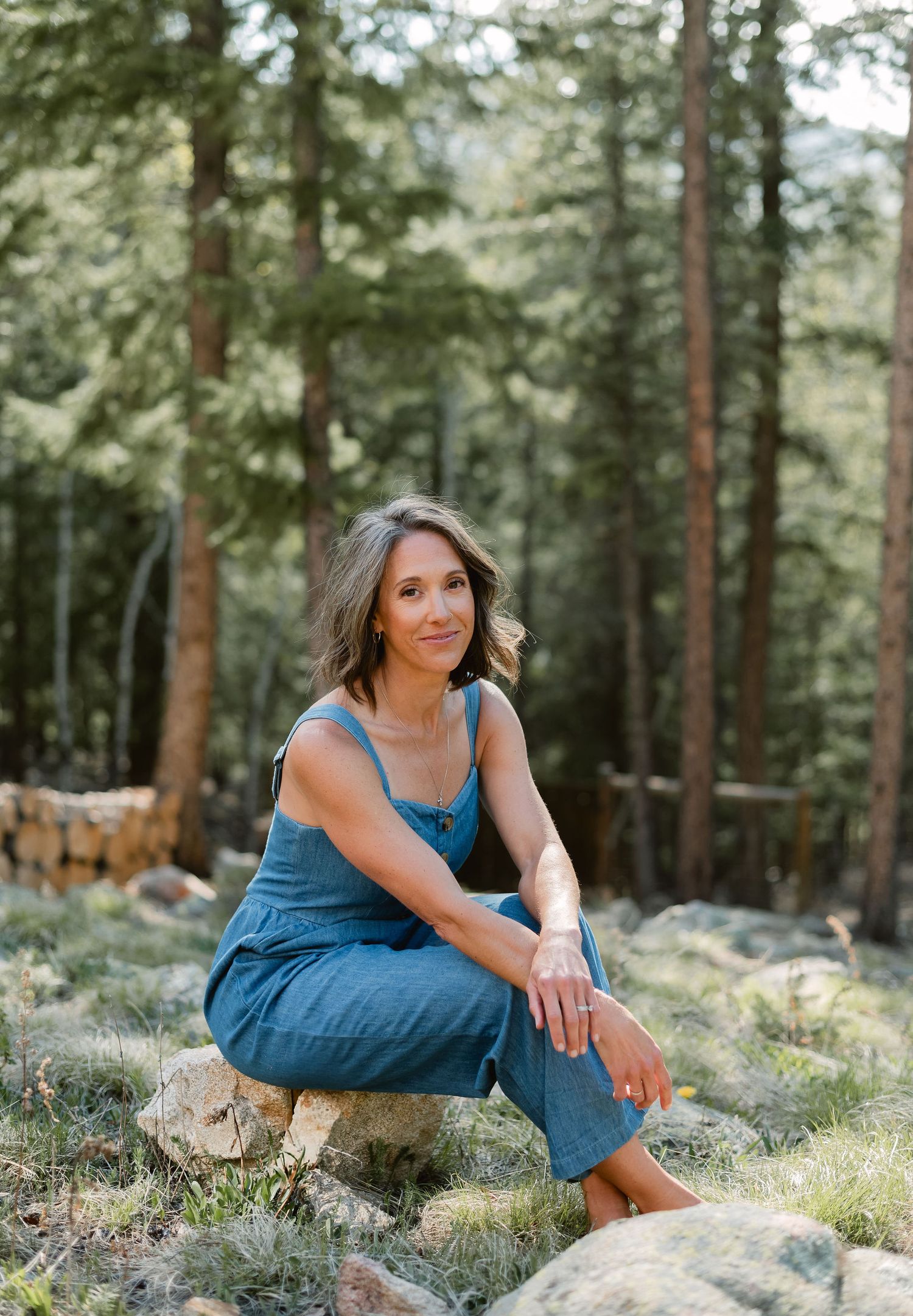 A woman in a blue dress sits gracefully on natural stone formations in a serene pine forest setting.