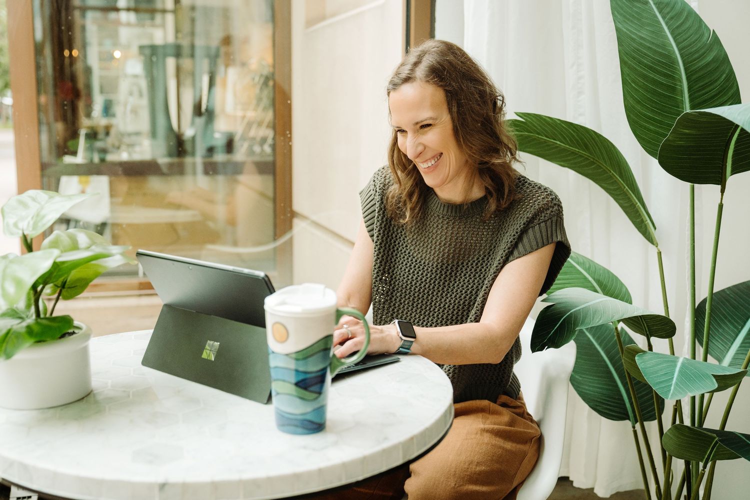 A person sits at a desk with a laptop and houseplants in a bright, modern workspace with natural lighting.