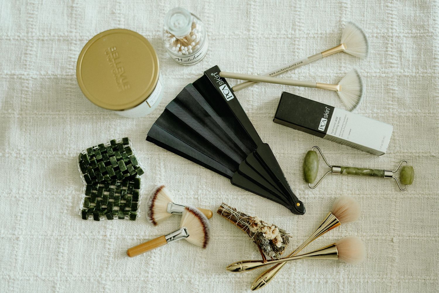 Flat lay of beauty tools including jade roller, fan brush, makeup brushes, hair clip, and compact on white textile.