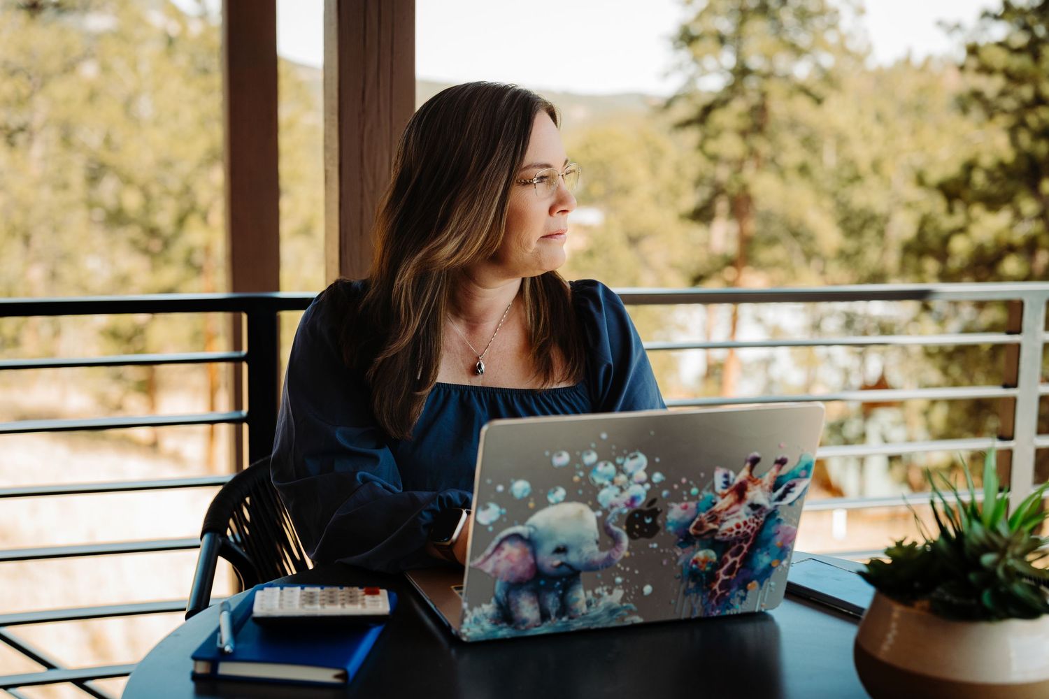 Woman smiling at laptop with animal sticker design, sitting at outdoor balcony desk surrounded by trees.