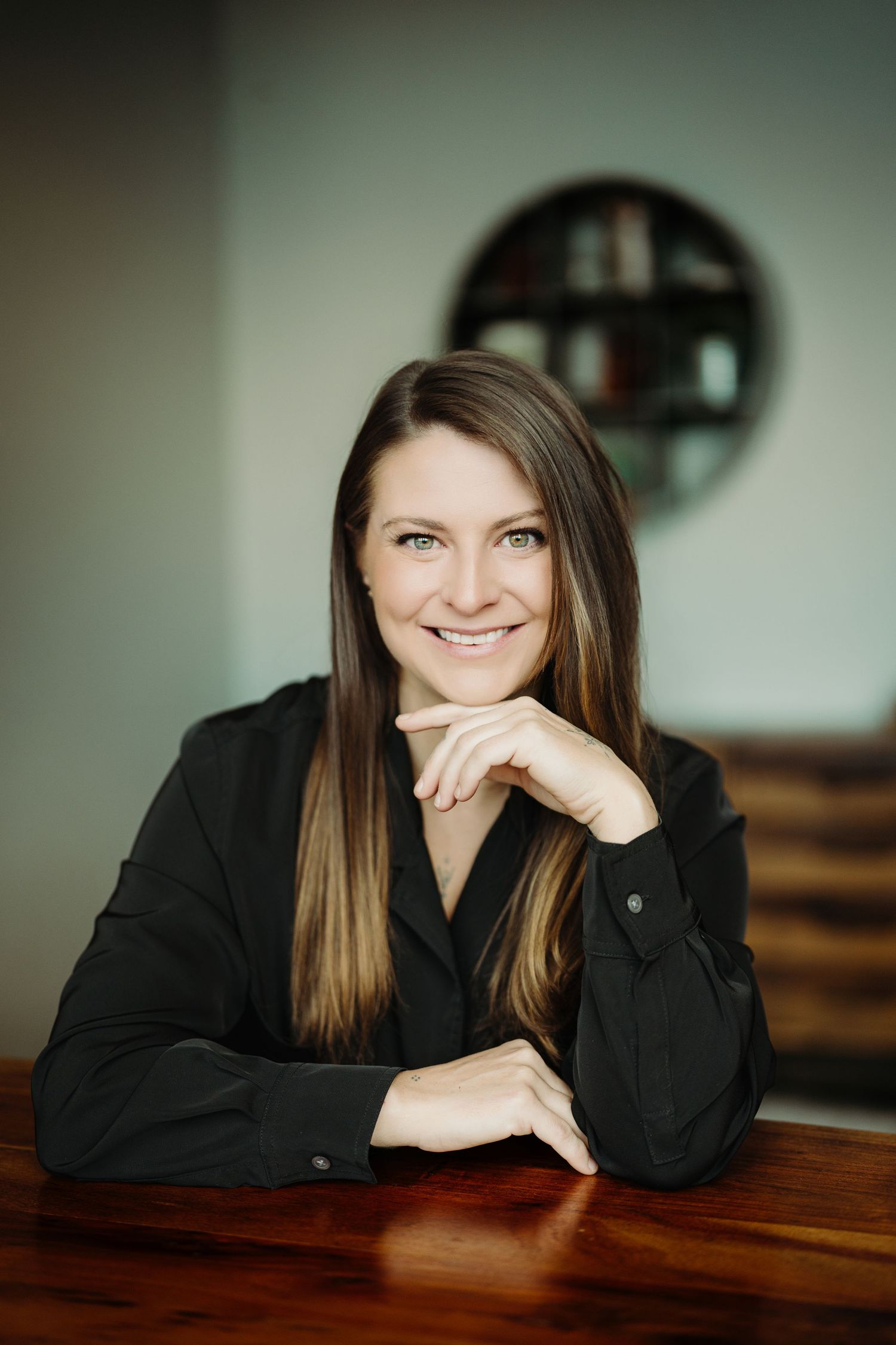 Smiling woman with long brown hair in black blazer, seated at wooden table with chin resting on hand.