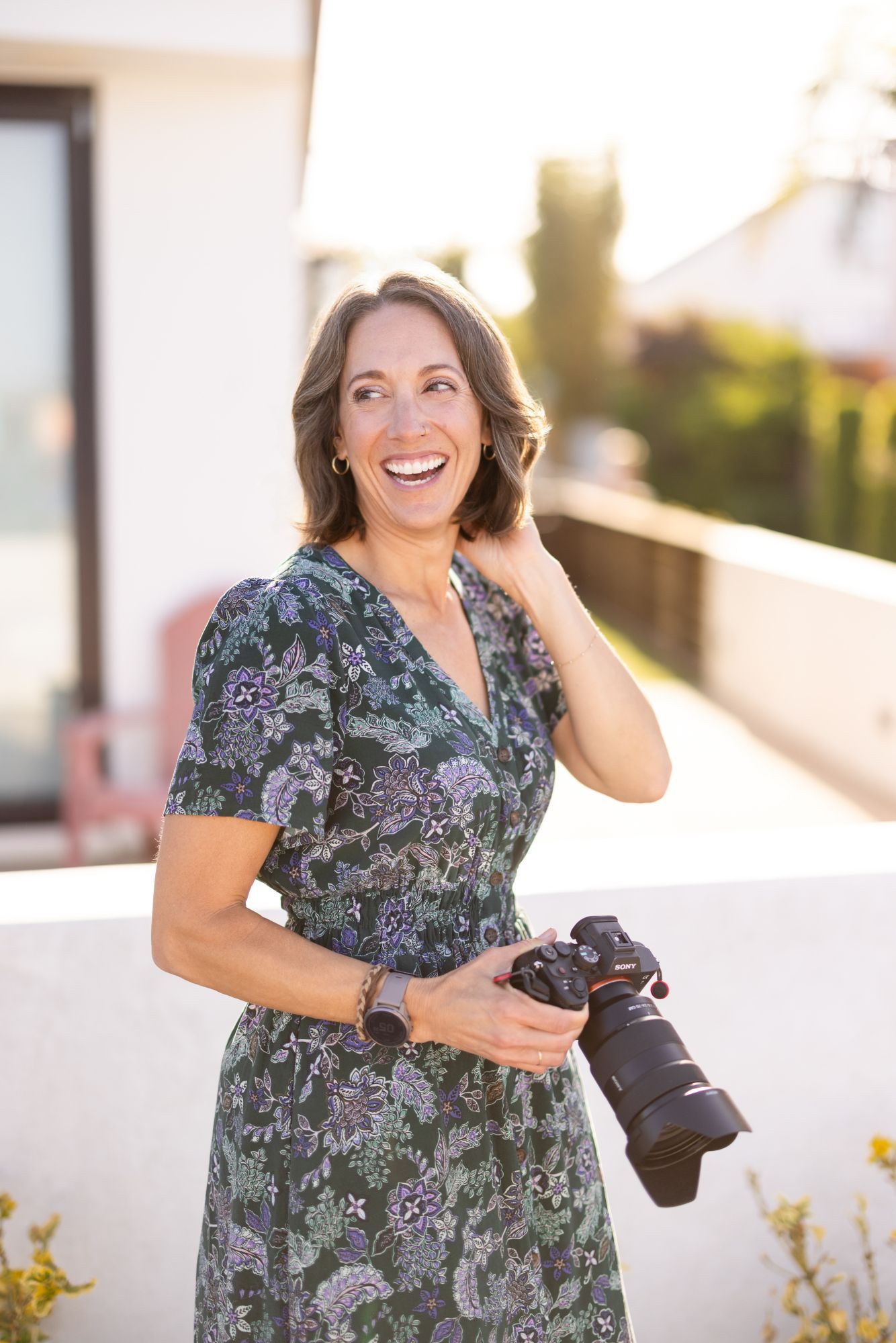 Smiling woman in floral dress holding a DSLR camera outdoors in warm sunlight, perfect for photography professional profiles.