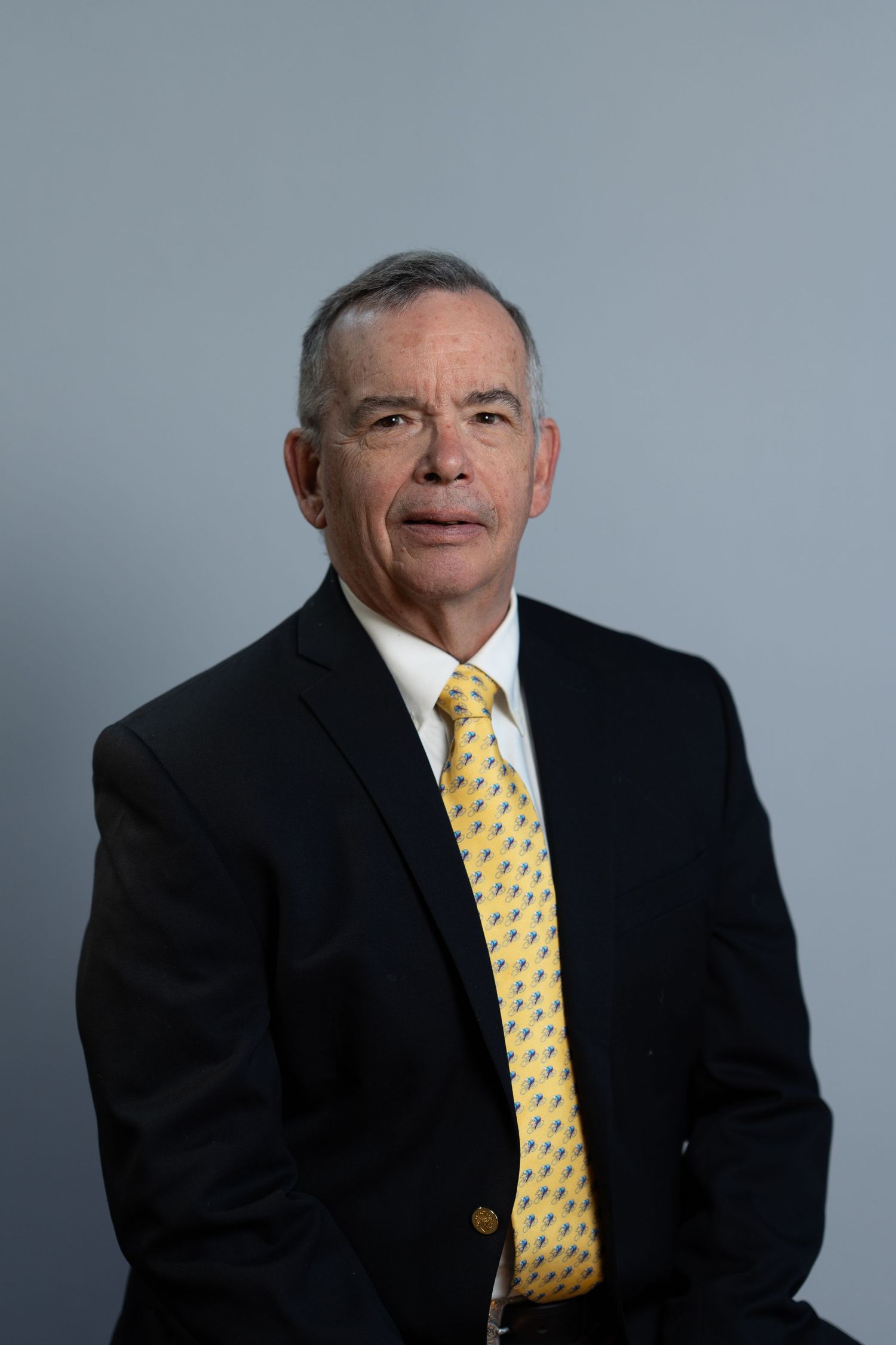 Professional headshot of a middle-aged man in a dark suit and yellow patterned tie against a gray background.