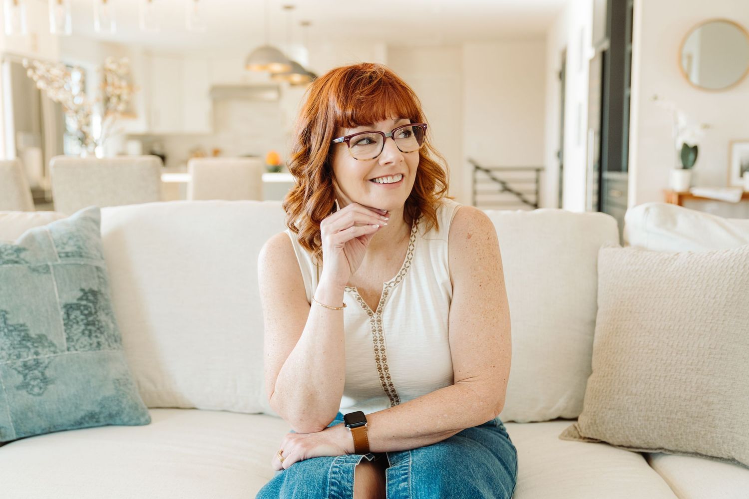 A smiling redheaded woman with glasses sits on a white sofa in a bright modern living room.