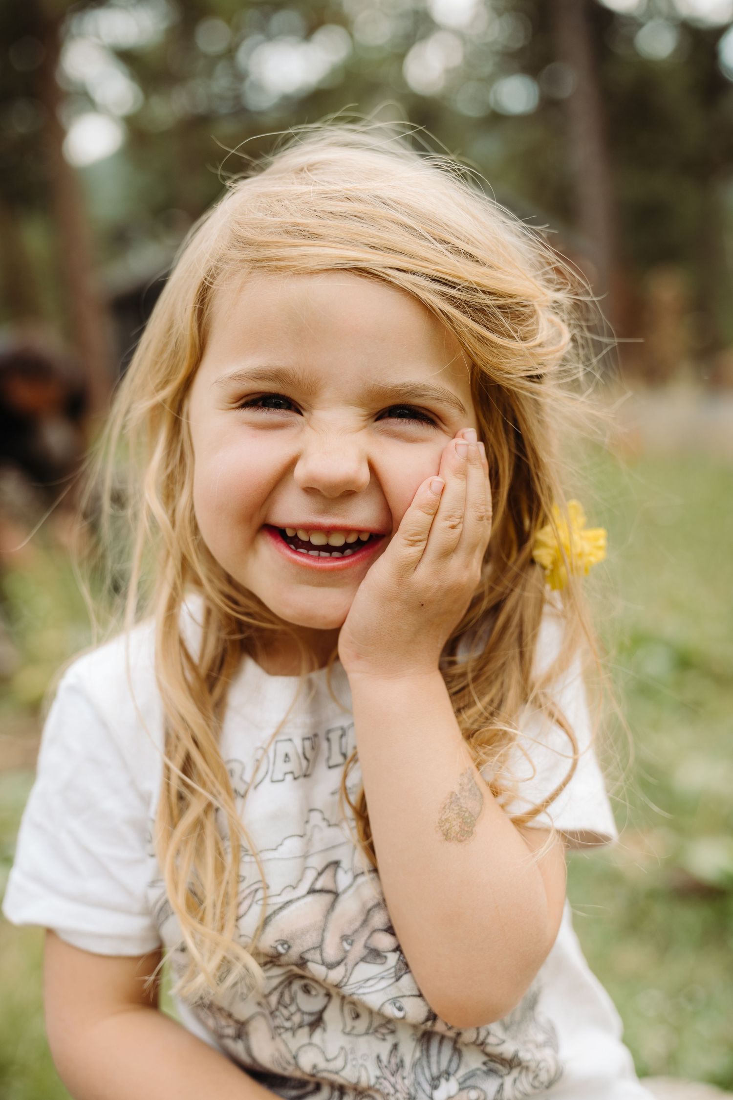 Smiling blonde little girl holding a yellow flower near her cheek outdoors in a forest setting.