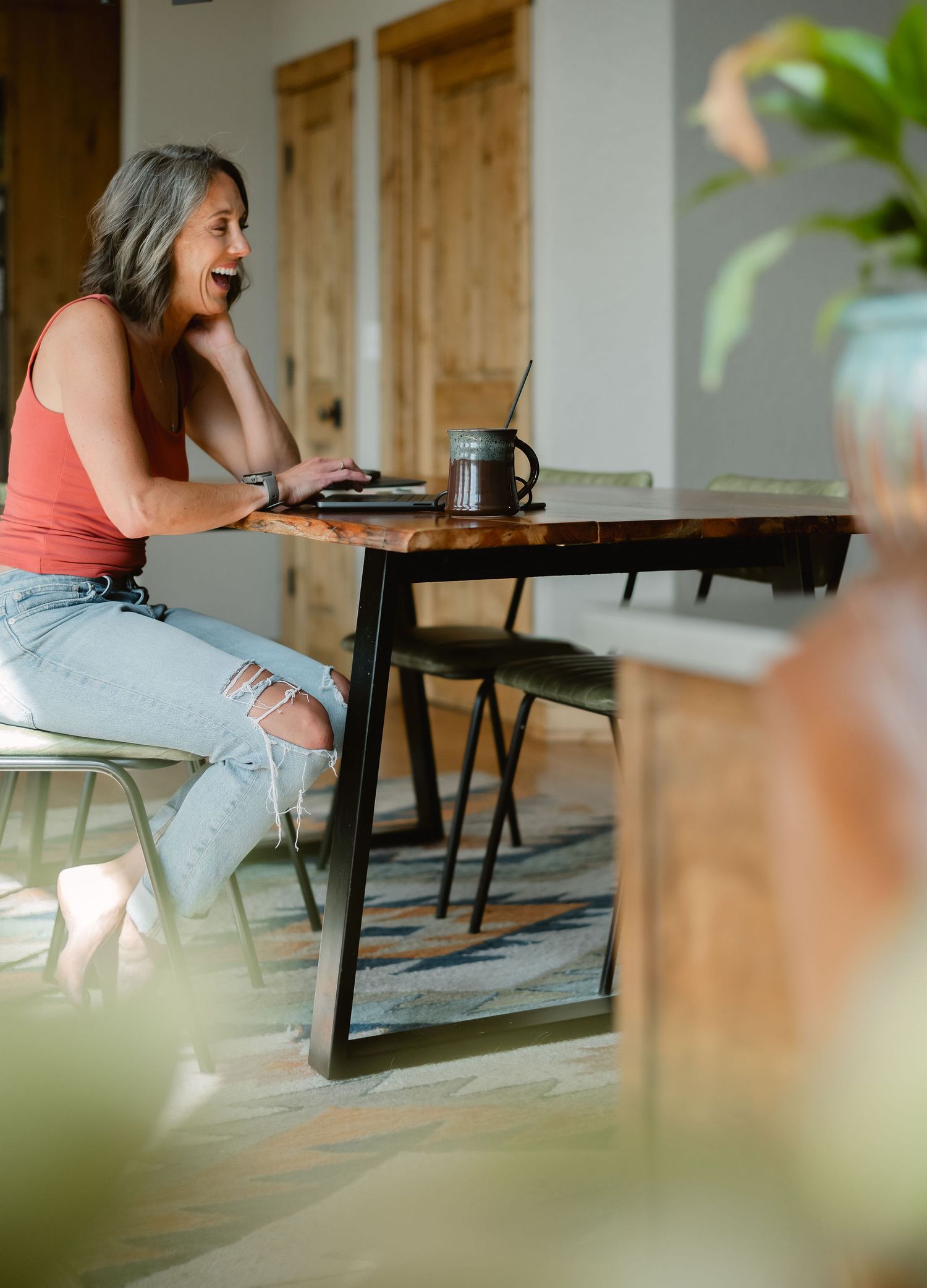 A woman with gray hair laughs while working on a laptop at a wooden table in a modern cafe.