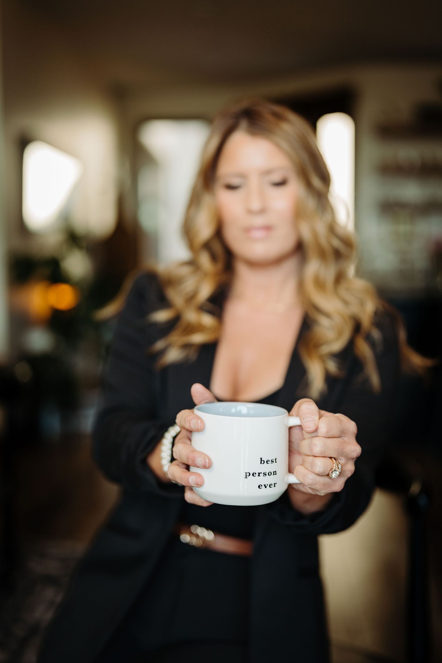 Woman in black blazer holding a white mug that reads best person ever, with blurred home interior background.