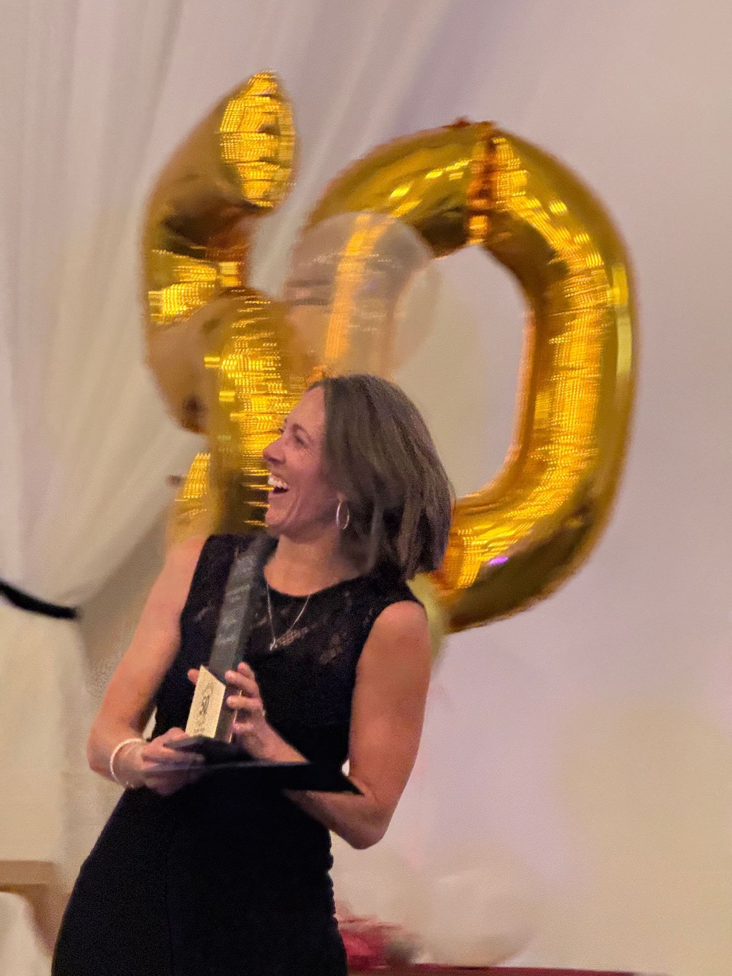 A speaker in a black dress celebrates in front of large golden number balloons at a special event.