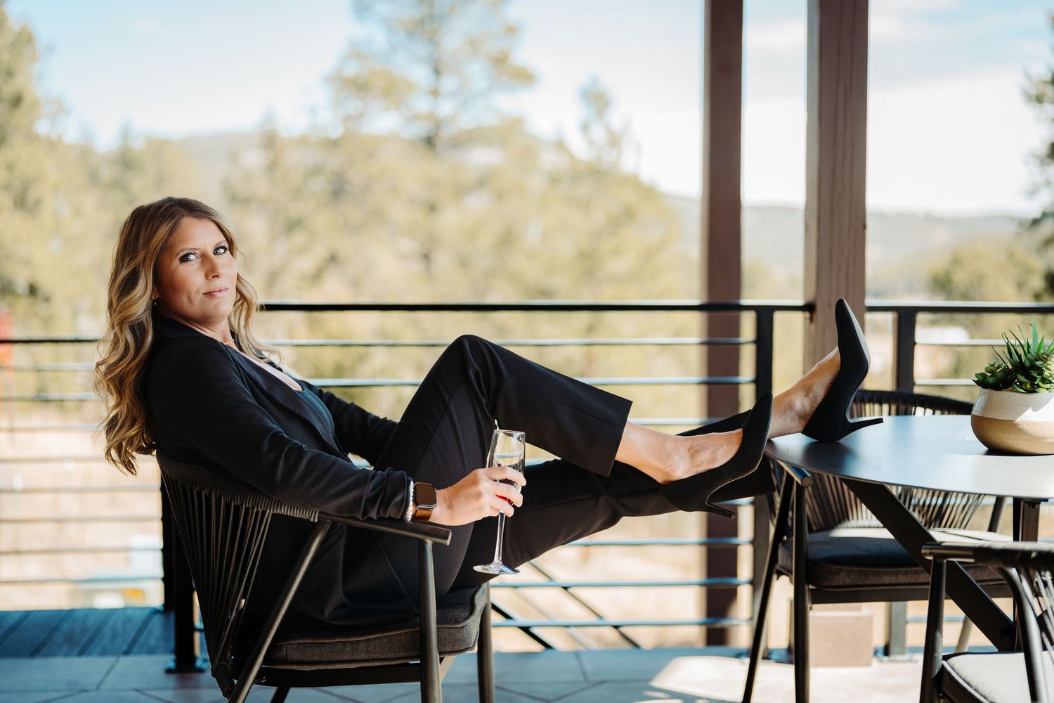 Woman in black suit relaxing on outdoor patio chair with wine glass, scenic forest view in background.