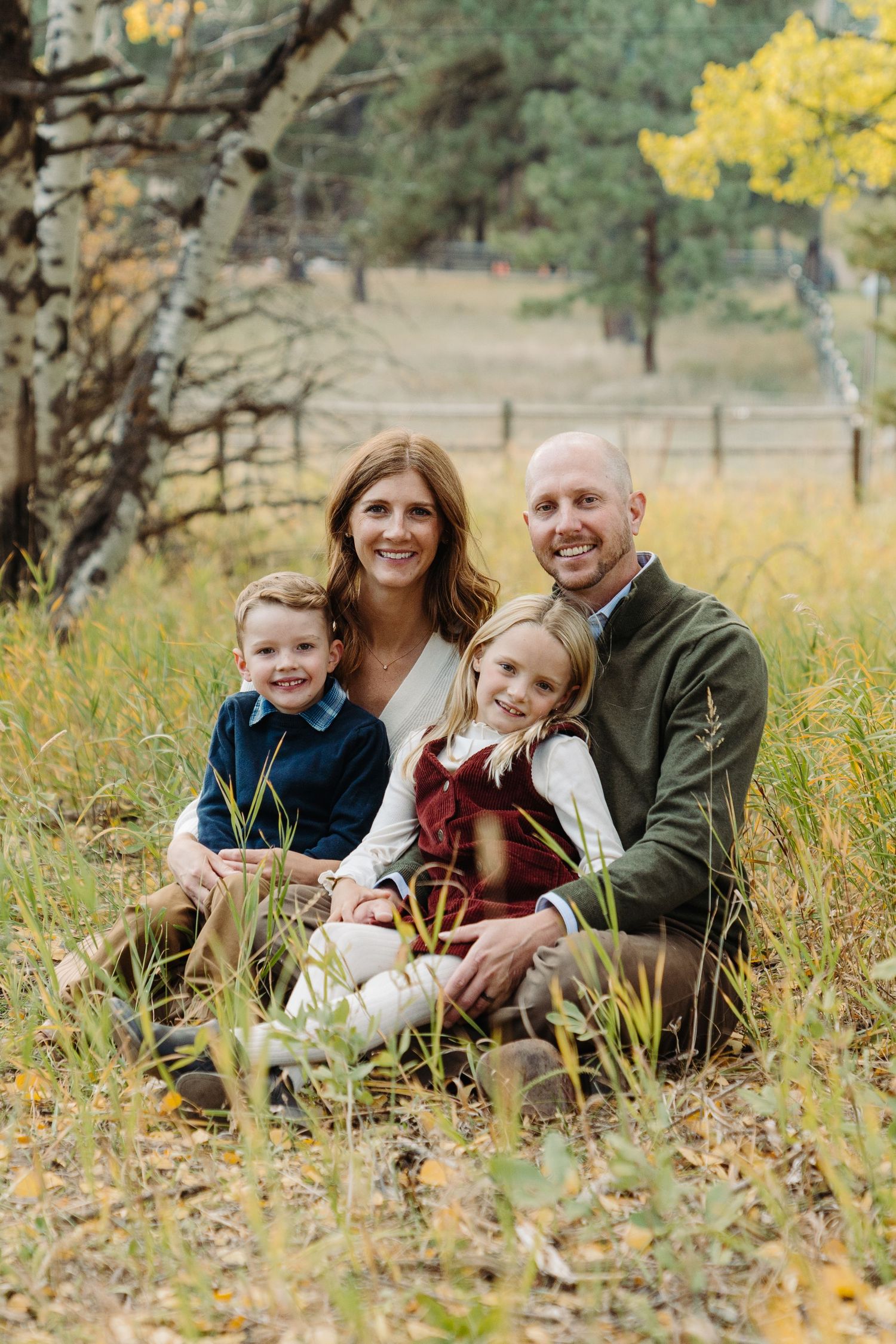 Family of four smiling outdoors in autumn meadow with colorful fall foliage, parents and two young children sitting in grass.