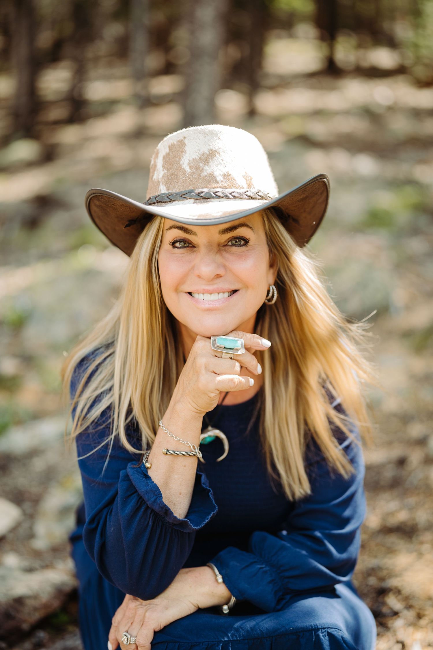 Portrait of someone in a navy sweater and light colored cowboy hat with silver jewelry sitting outdoors in nature.