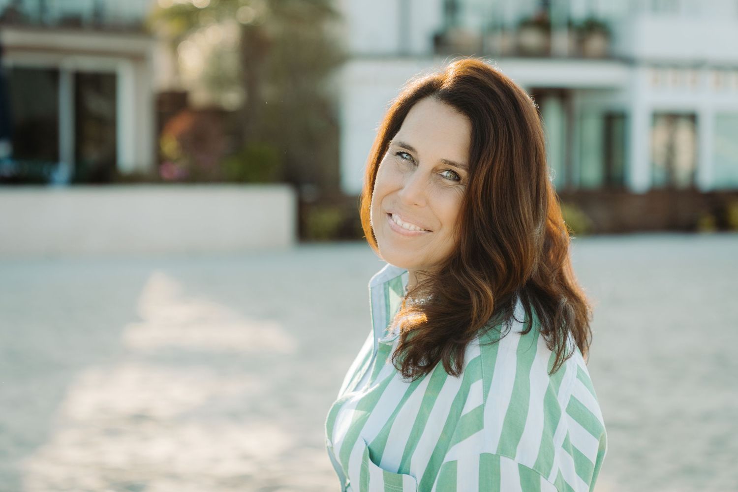 Smiling brunette woman in green striped shirt outdoors in golden sunlight near white building.