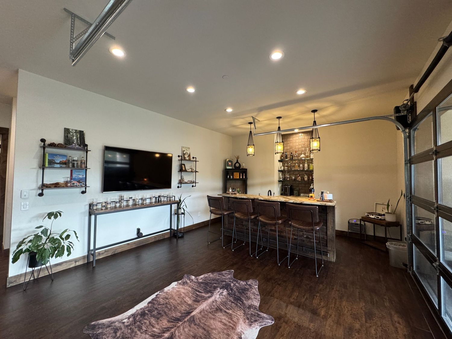 Modern home bar area with dark wood floors, industrial shelving, and mounted TV features sleek black barstools and recessed lighting.