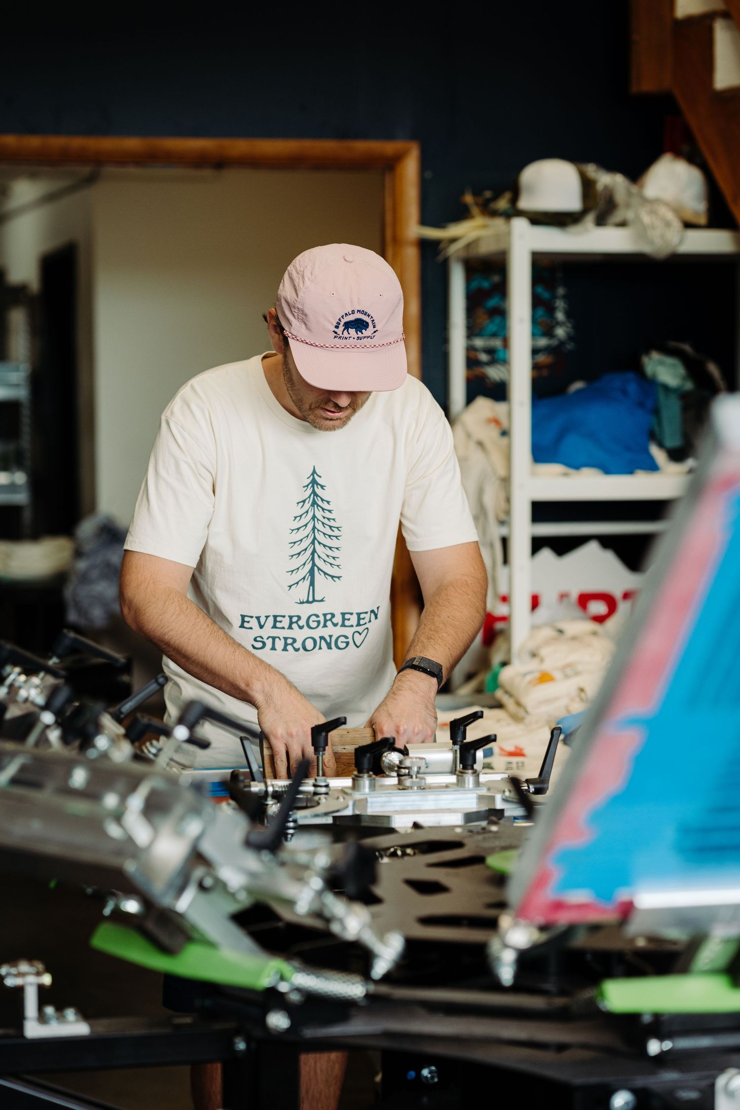 Person wearing pink cap and Evergreen Strong shirt works on ski or snowboard maintenance at repair shop workbench.
