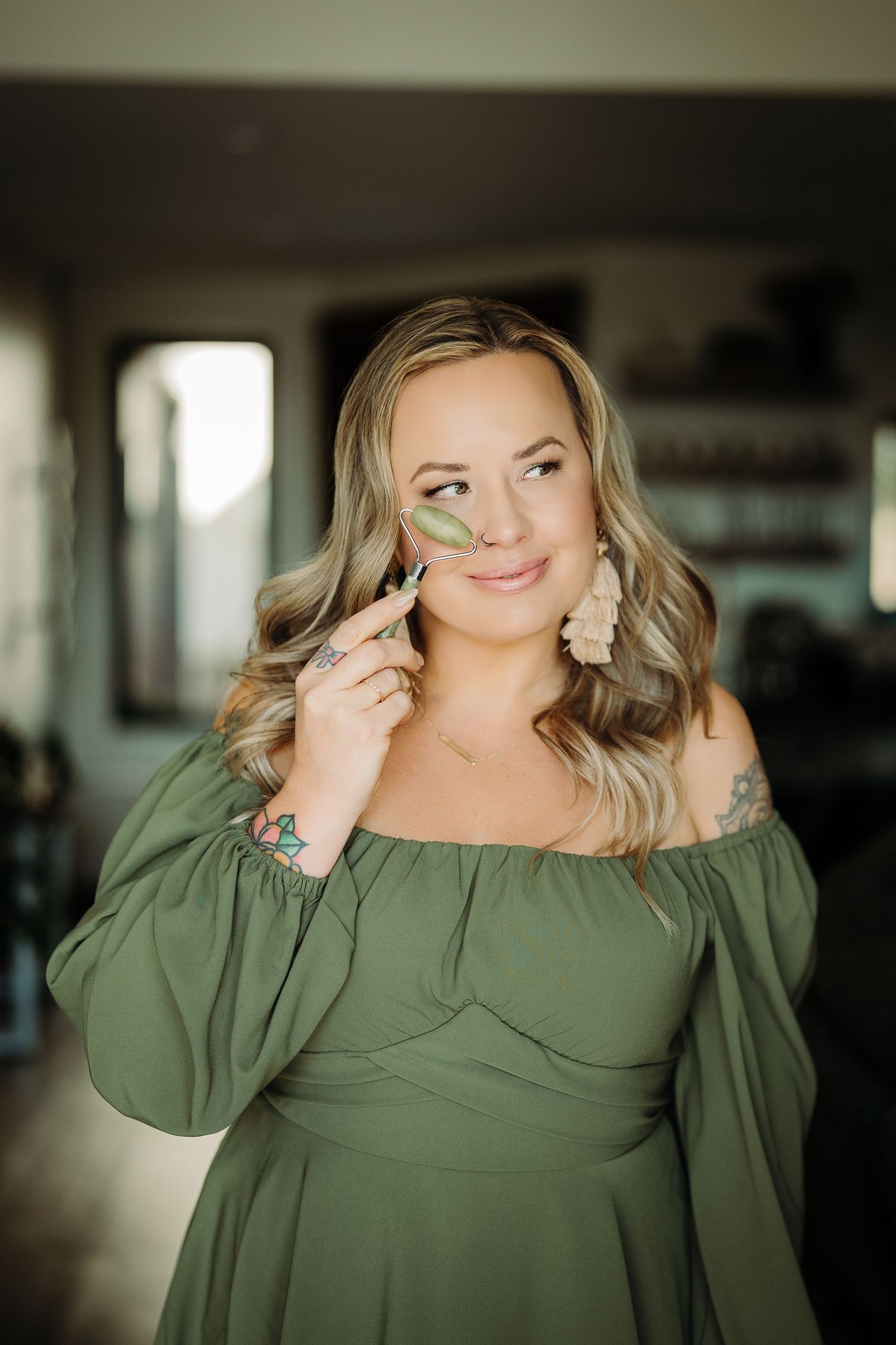 Woman in green off-shoulder dress holds jade roller to cheek, smiling softly indoors.