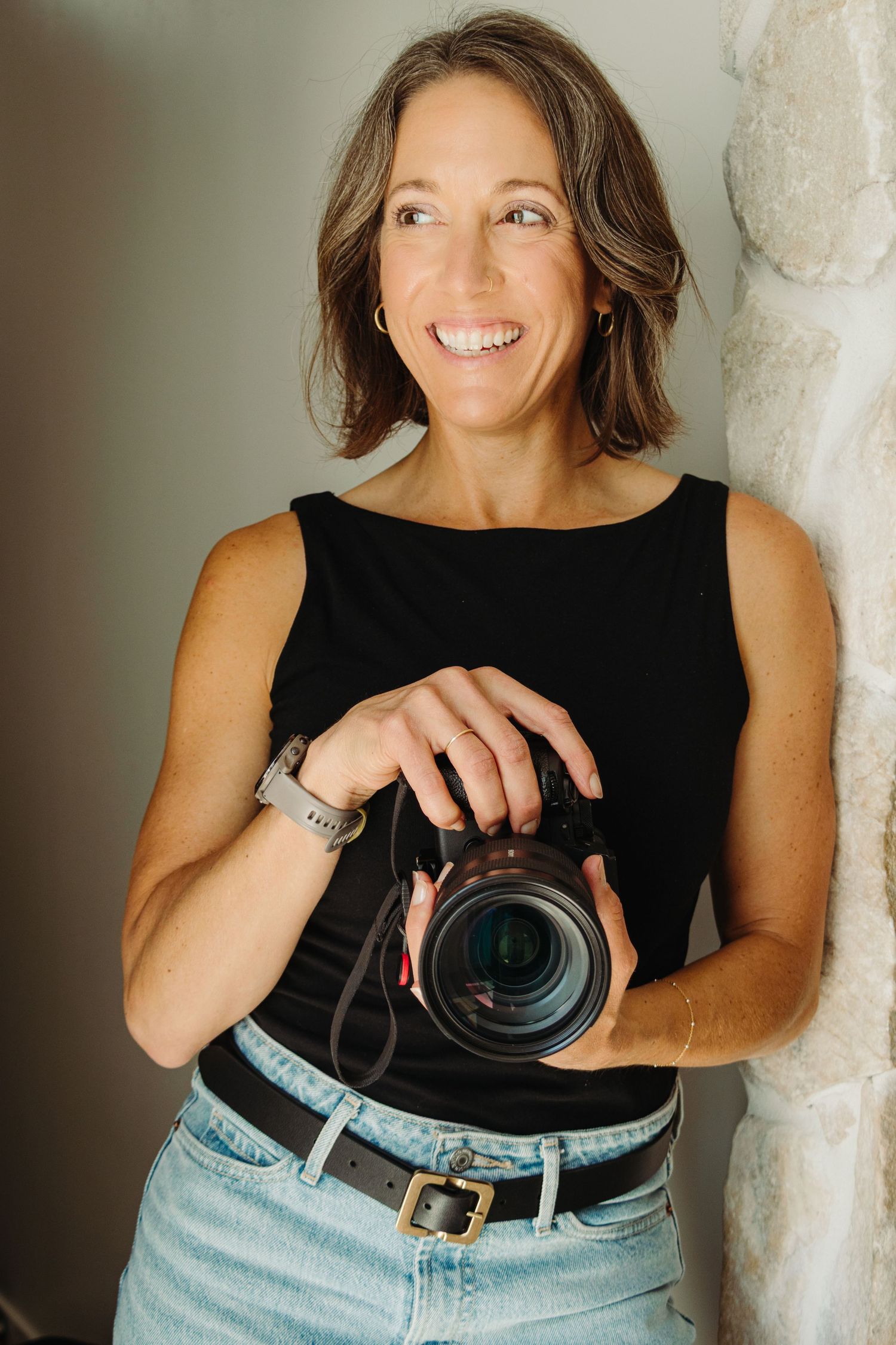 Smiling woman in black sleeveless top holding a professional DSLR camera, leaning against a stone wall.