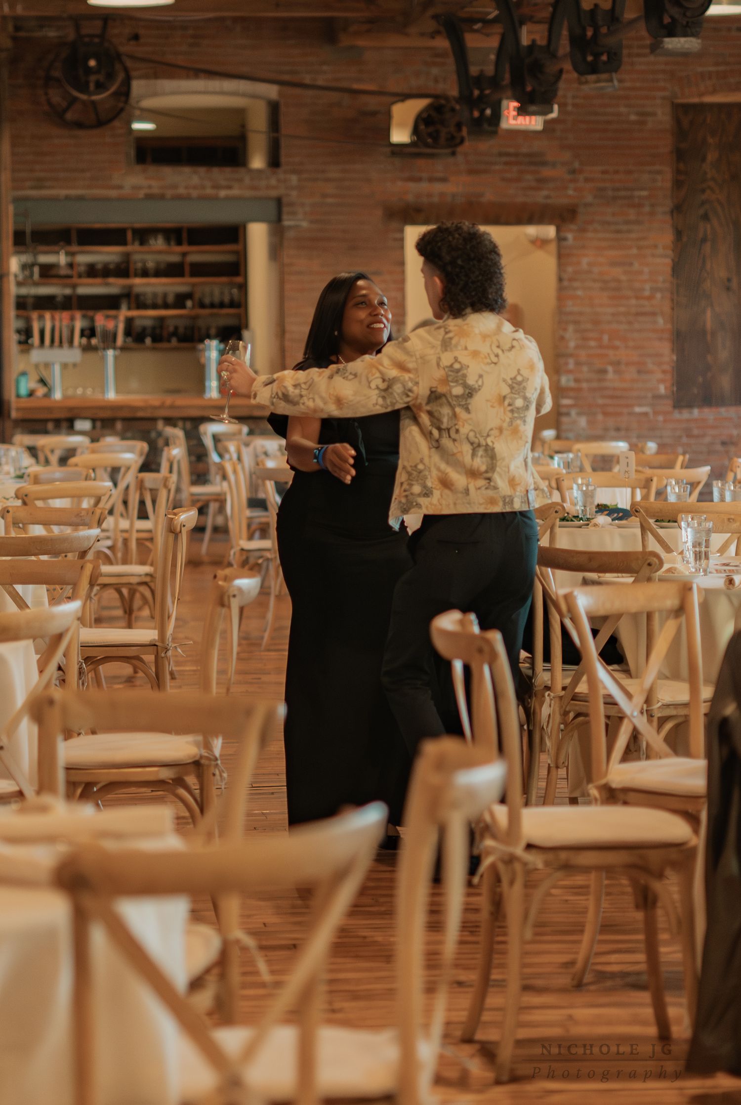 Two people dancing joyfully in an elegant rustic wedding venue with wooden chairs and brick walls.