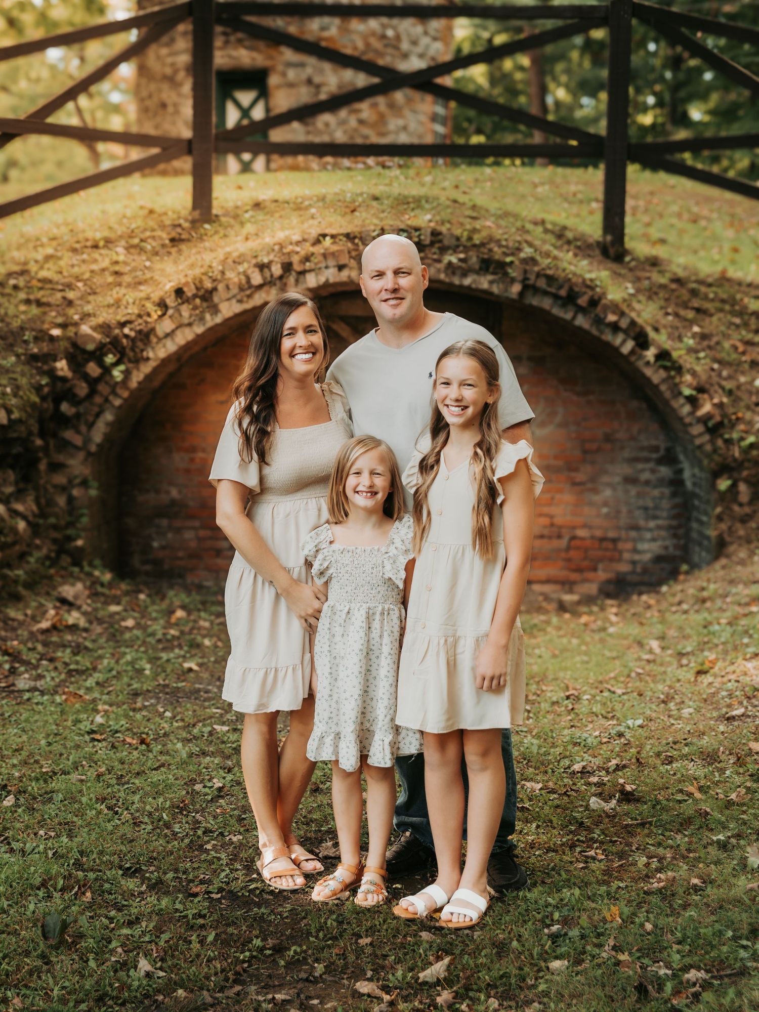 Family of four posing outdoors near a rustic brick arch bridge, wearing coordinating neutral outfits in a lush green setting.