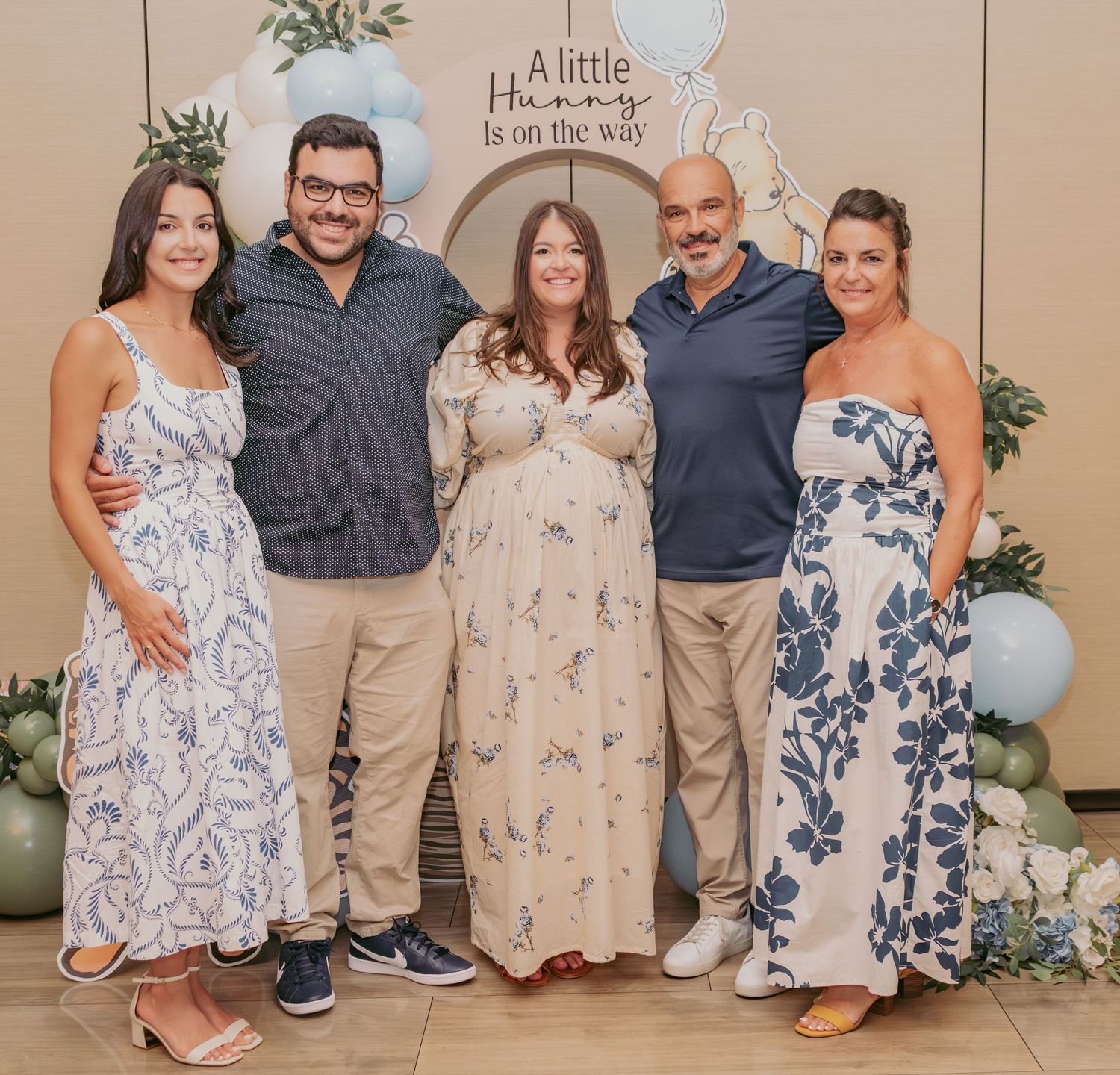Five people pose together at a baby shower with balloon decorations and a Winnie the Pooh themed sign behind them.