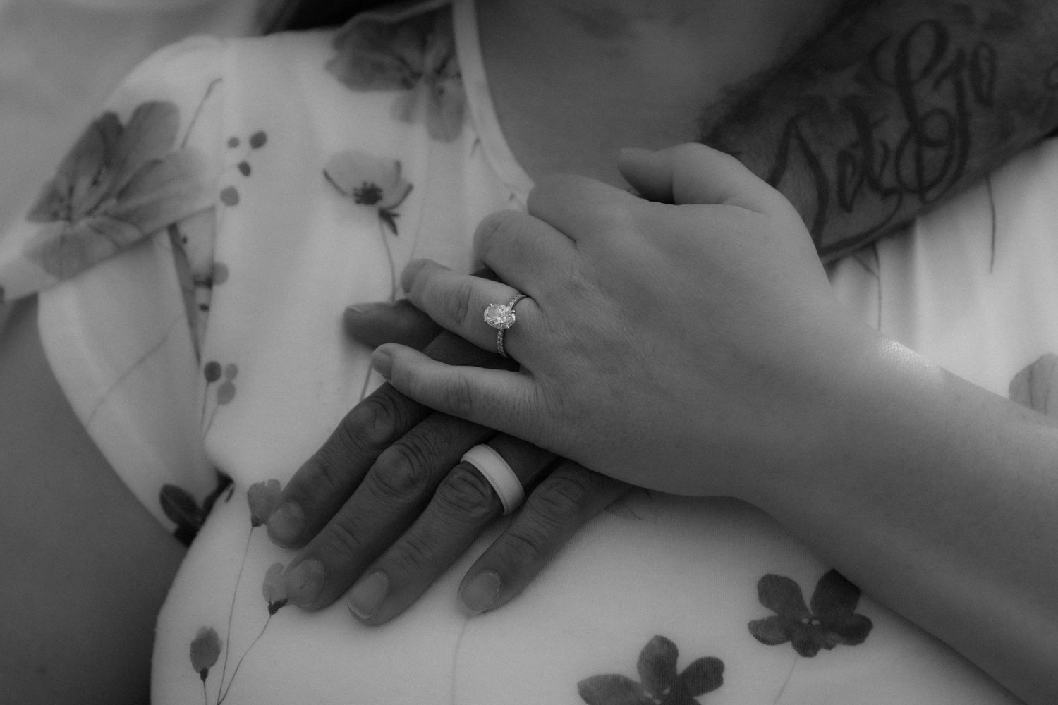 Black and white photo of couple's hands with diamond engagement ring, floral dress and tattoos visible.