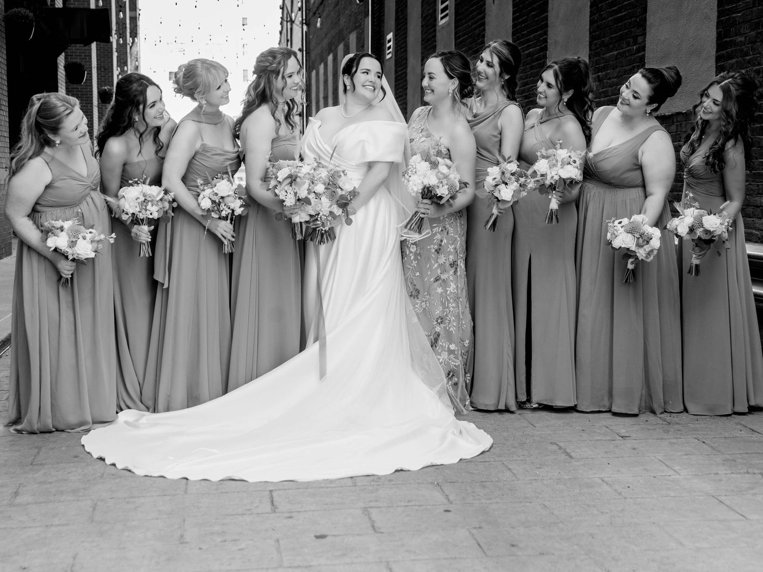 Bride in white gown poses with bridesmaids in sage green dresses holding bouquets in black and white photo.
