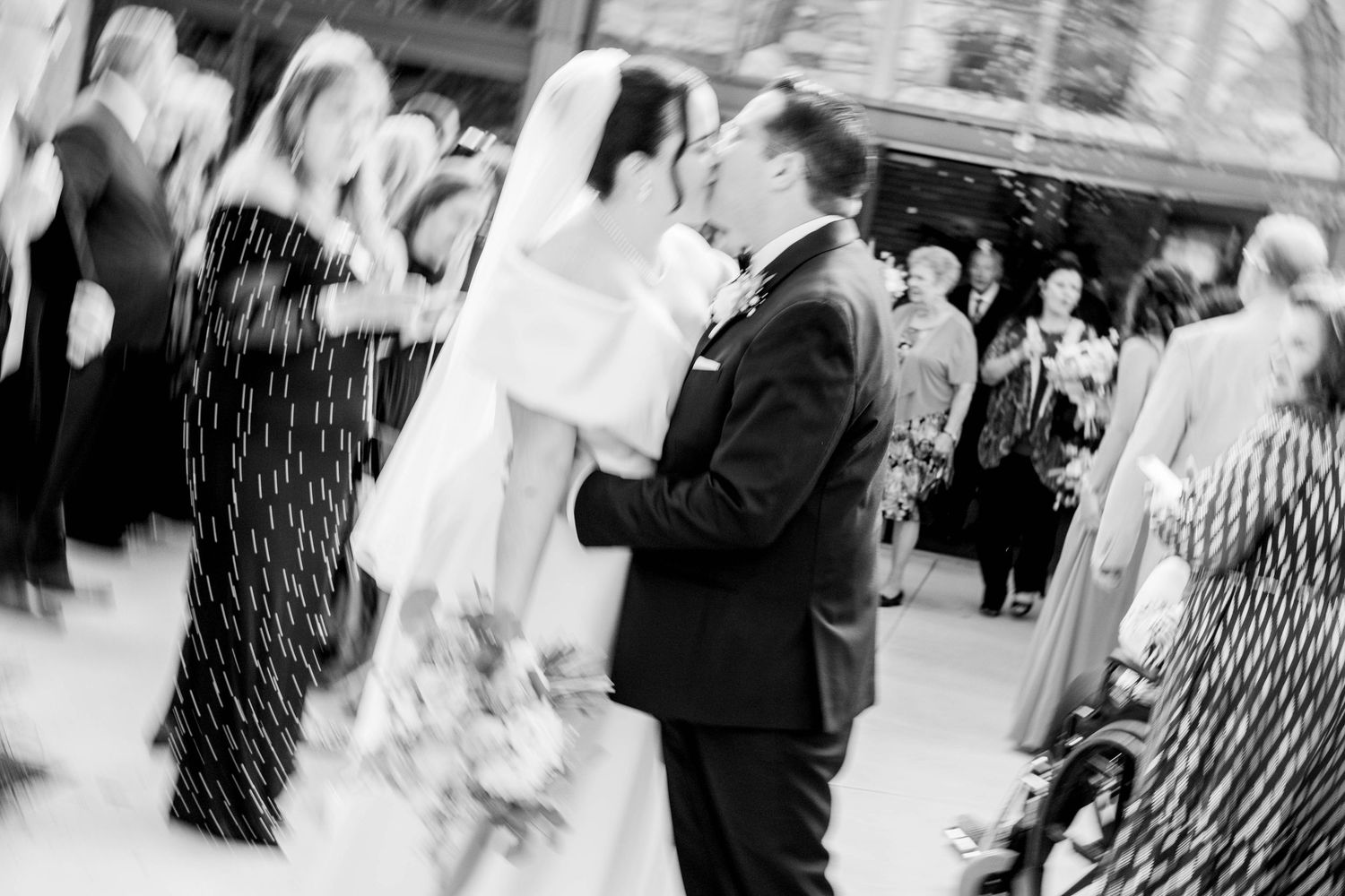 Black and white photo of a bride and groom sharing a kiss while guests celebrate around them at an outdoor wedding.