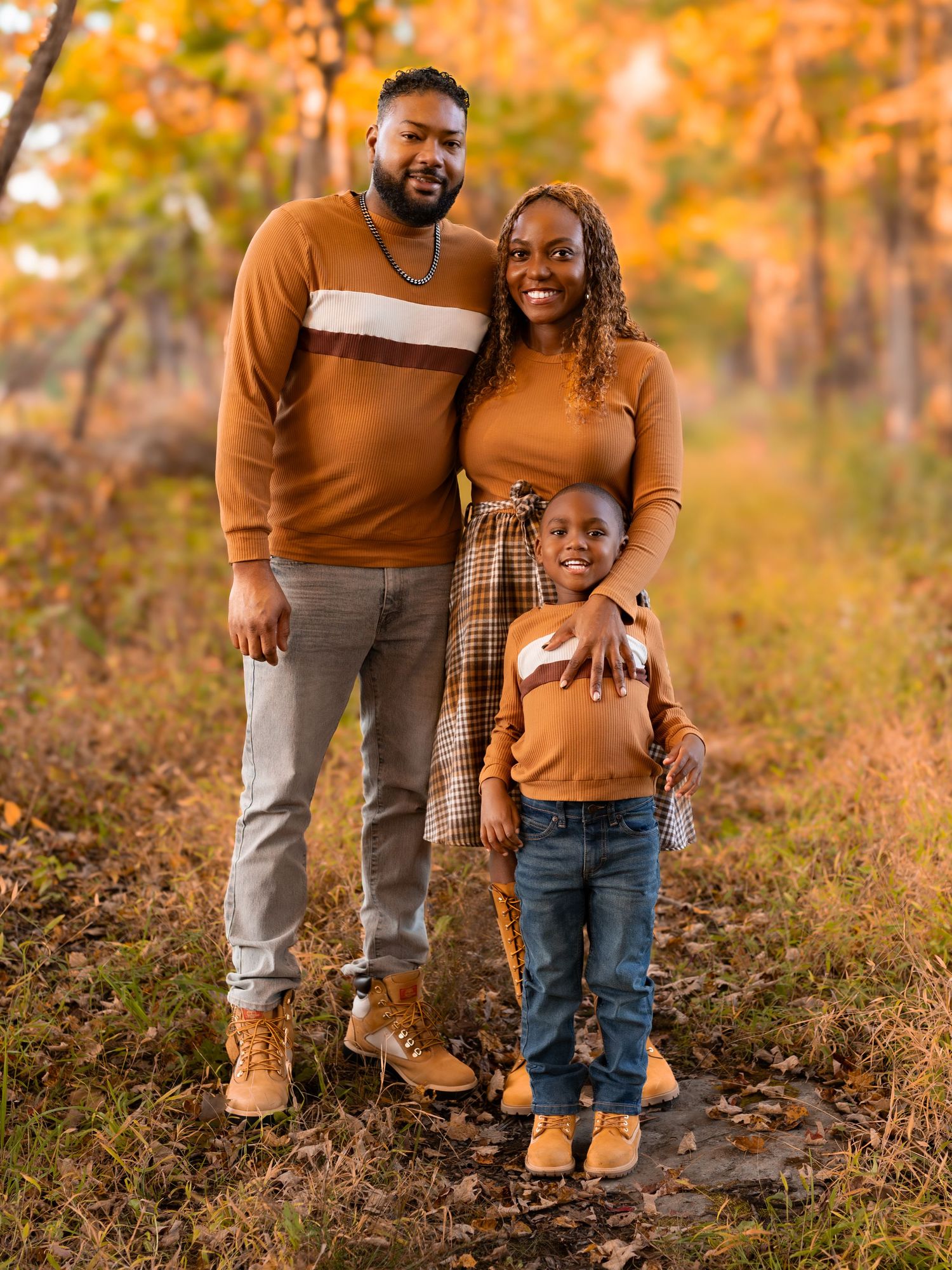 A family of three poses outdoors in autumn, wearing coordinated brown outfits among colorful fall foliage.