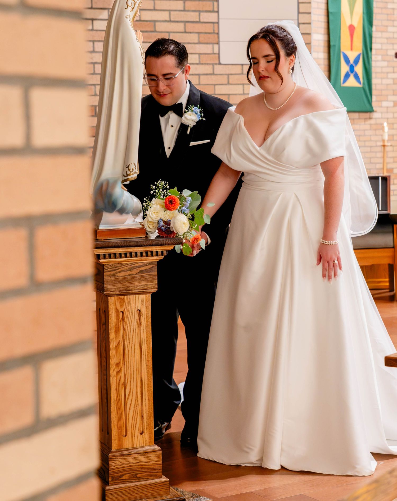 Bride in white off-shoulder gown and groom in black tuxedo exchanging vows at church altar.