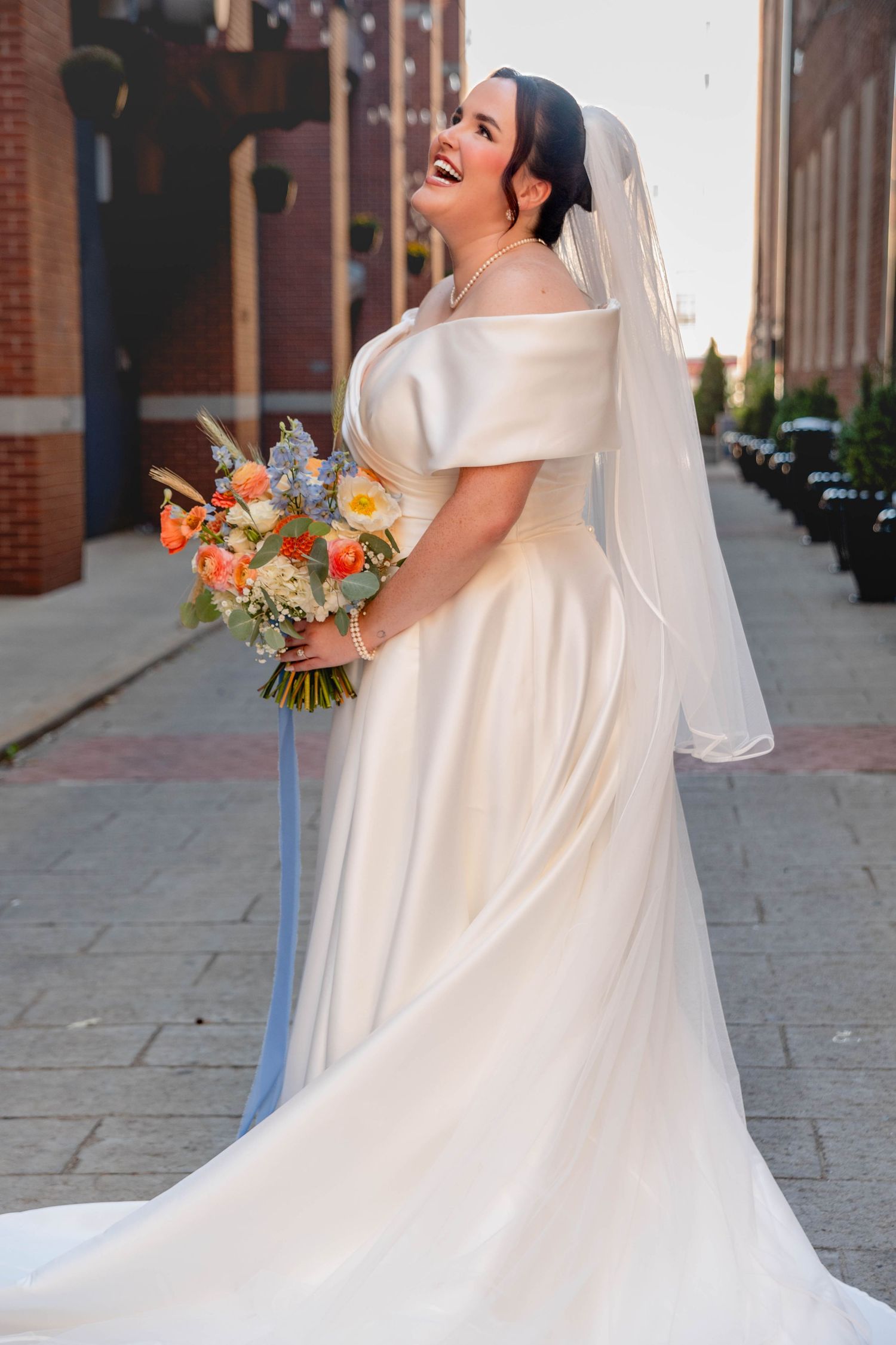 Plus-size bride in an off-shoulder white gown holding a colorful bouquet, smiling outdoors.