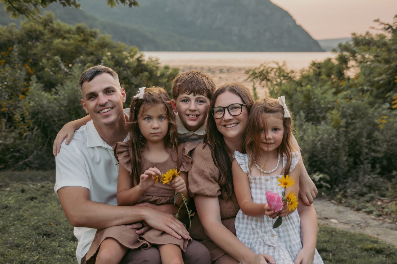 Happy family of five posing outdoors near a scenic mountain and river backdrop during golden hour.
