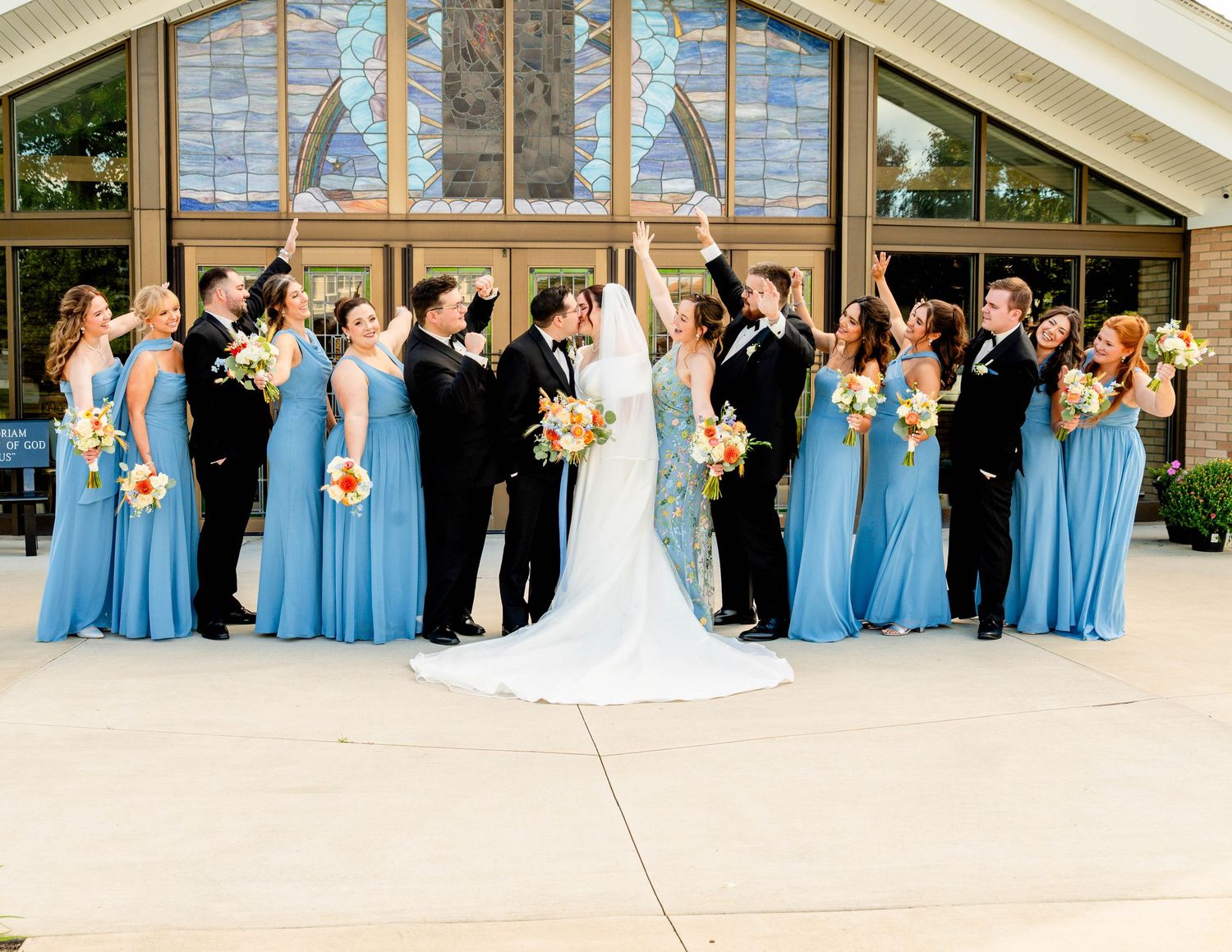 Wedding party in blue dresses celebrating outside church with bride and groom kissing.