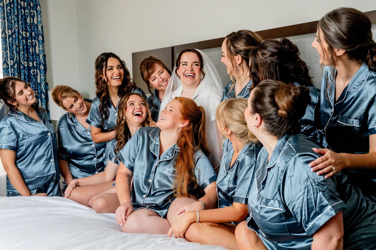 Bridesmaids in matching blue satin pajamas laughing and bonding with bride on bed during wedding morning preparations.