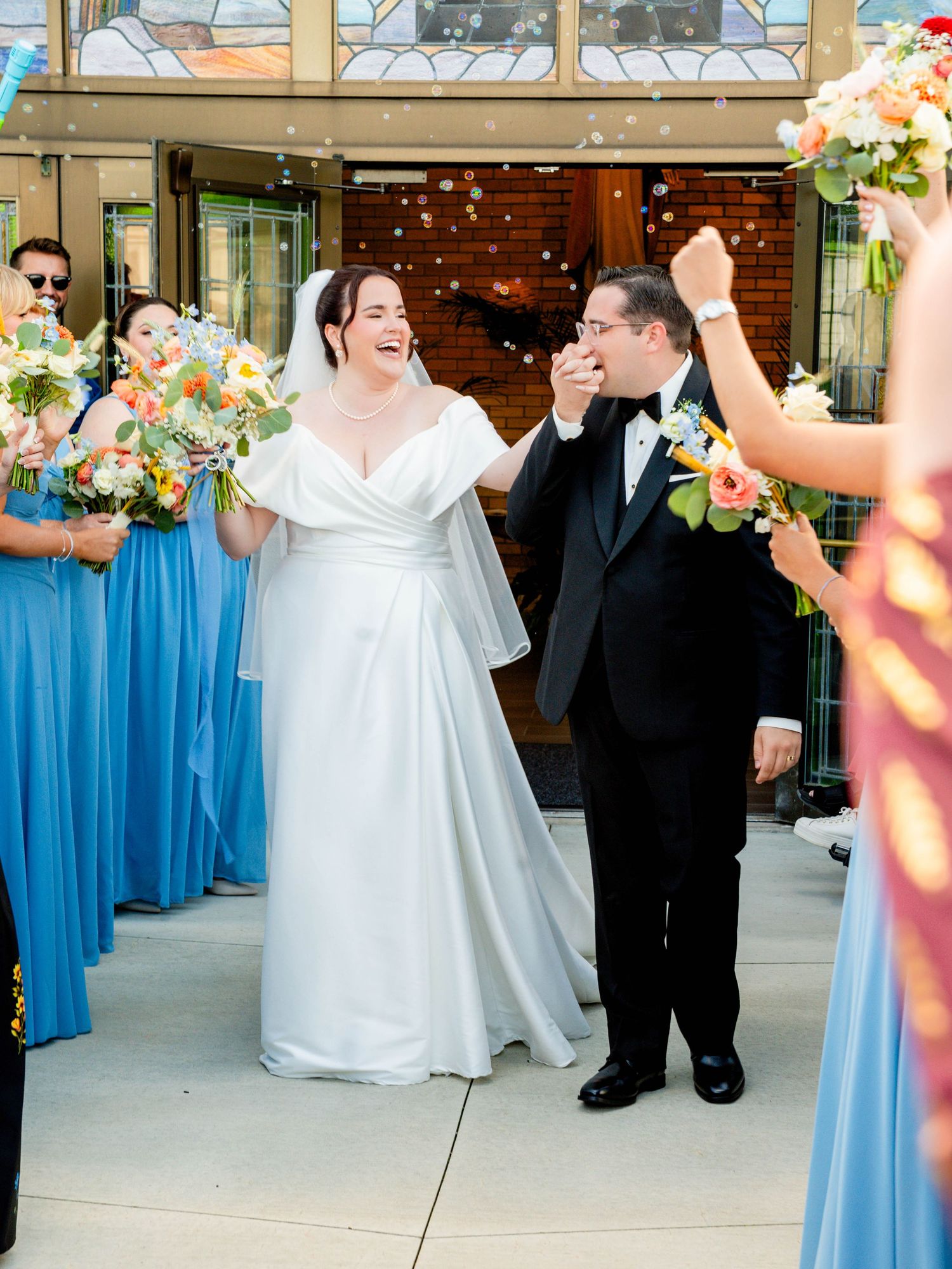Joyful newlywed couple exits church ceremony surrounded by bridesmaids in blue dresses as confetti fills the air.