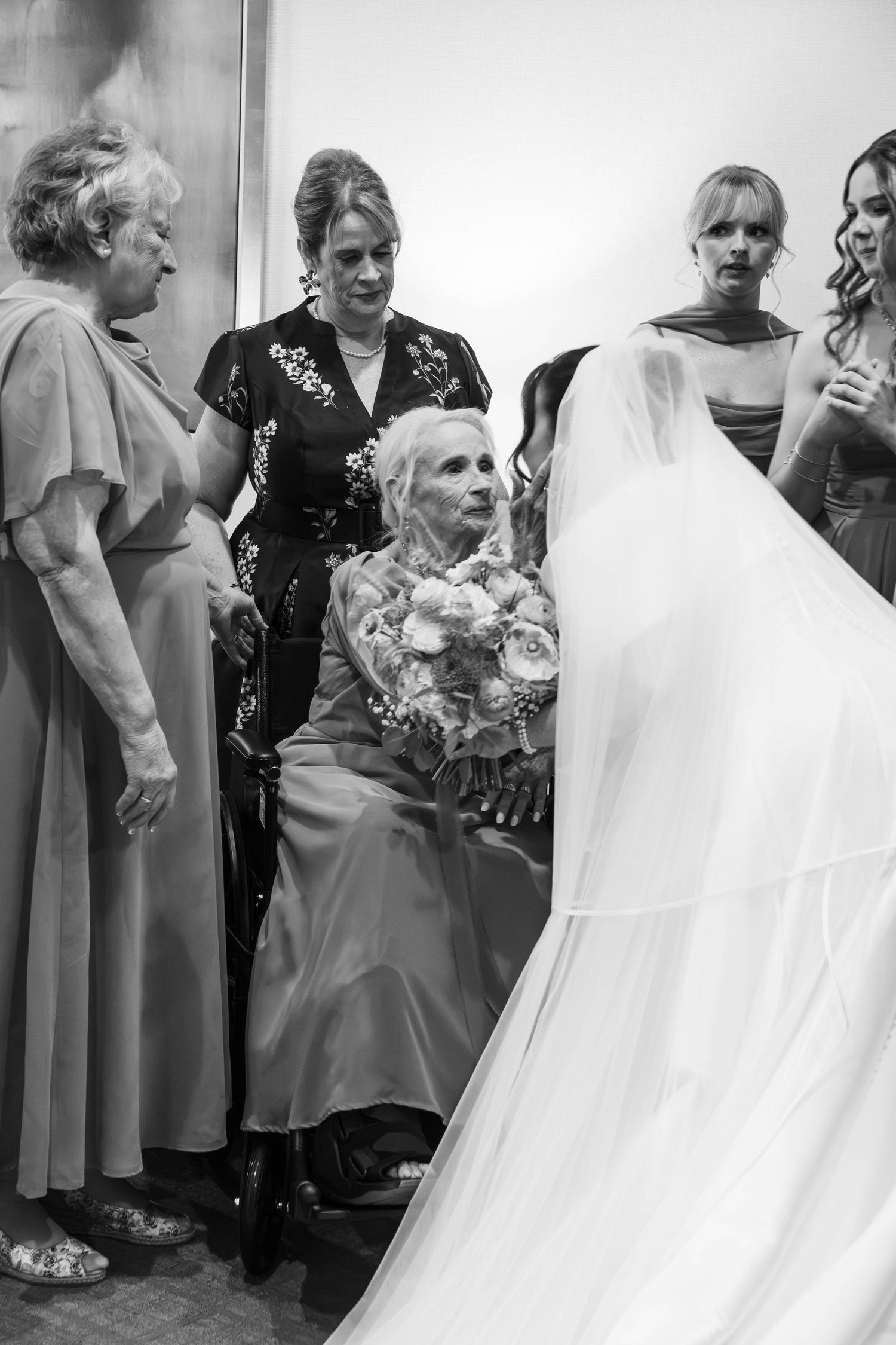 Bride in white gown and veil showing her bouquet to elderly seated woman surrounded by family in black and white photo.