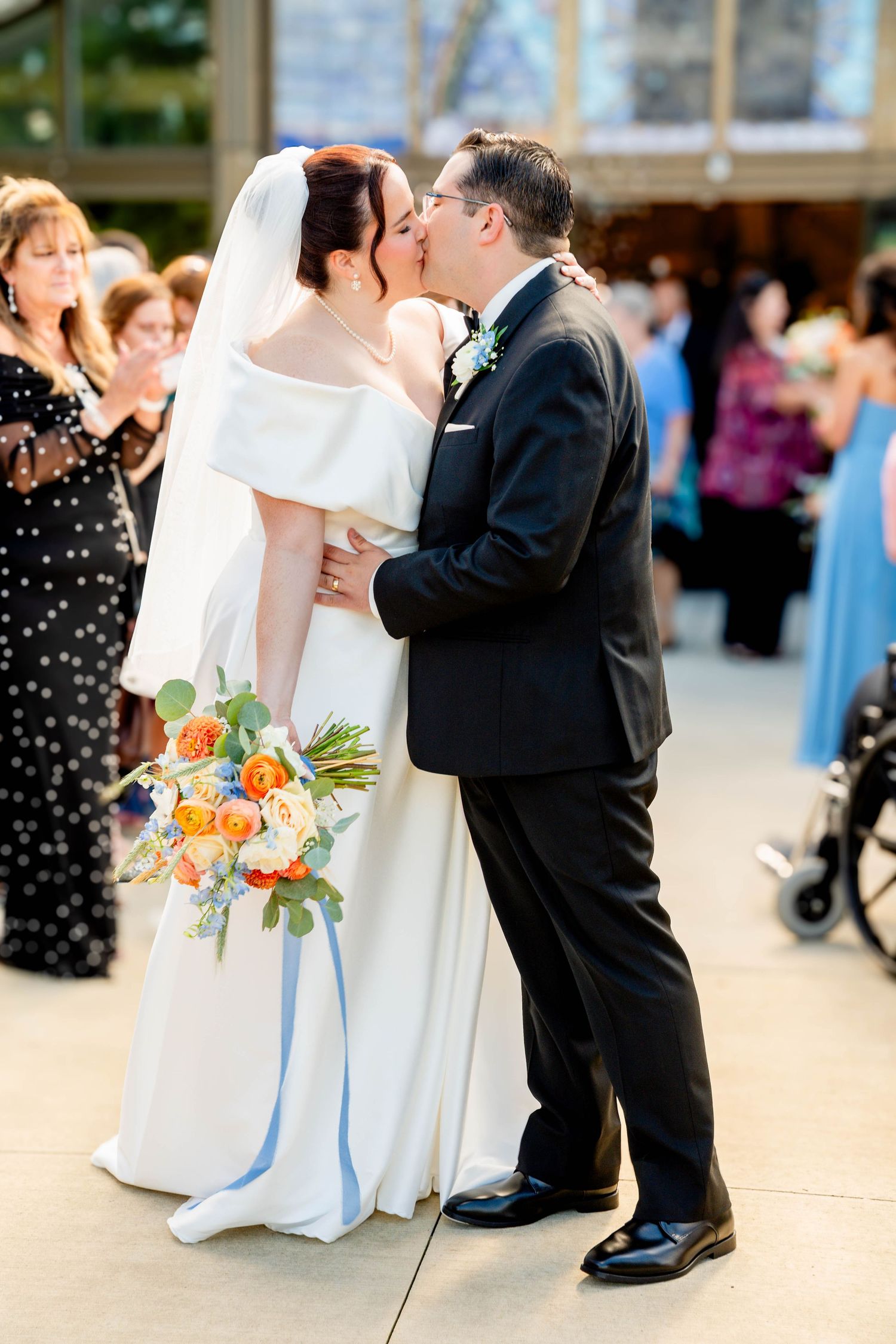 Bride and groom share first kiss at wedding ceremony, bride holding colorful orange and blue bouquet.