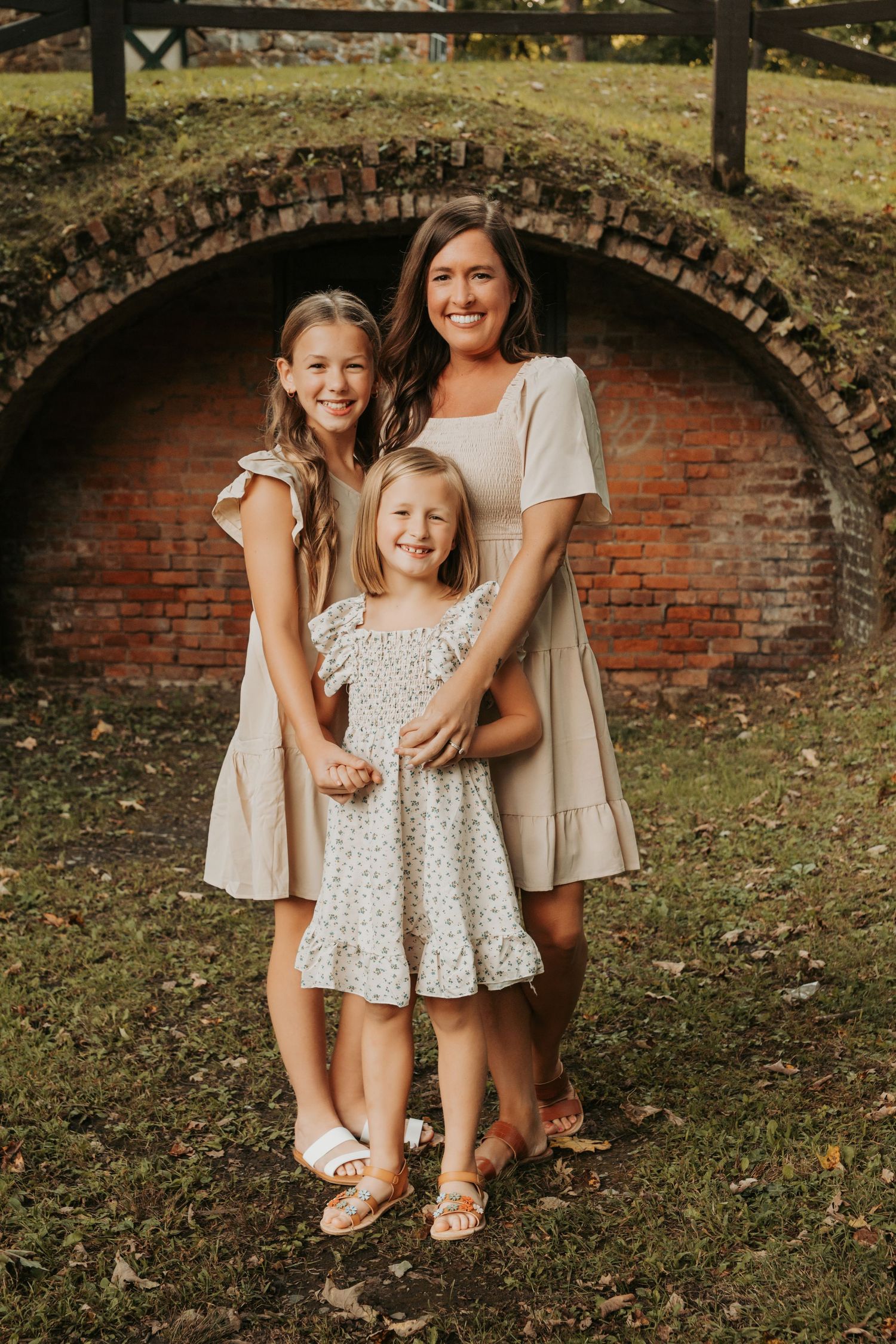 A mother and two daughters smile together in matching beige dresses before a rustic brick arch outdoors.
