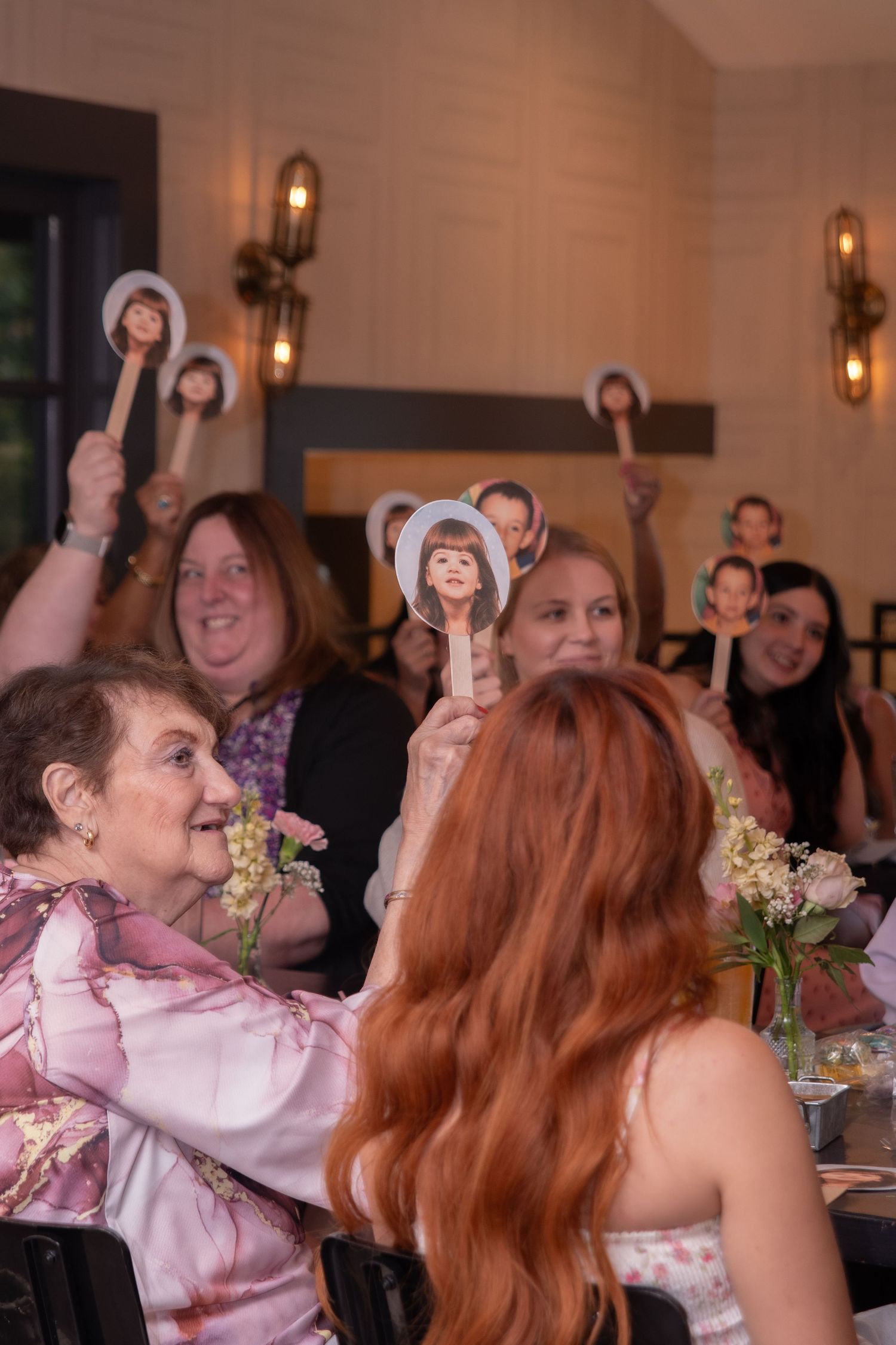 Guests at a celebration hold up face fans of the guest of honor, smiling and laughing at a decorated banquet table.