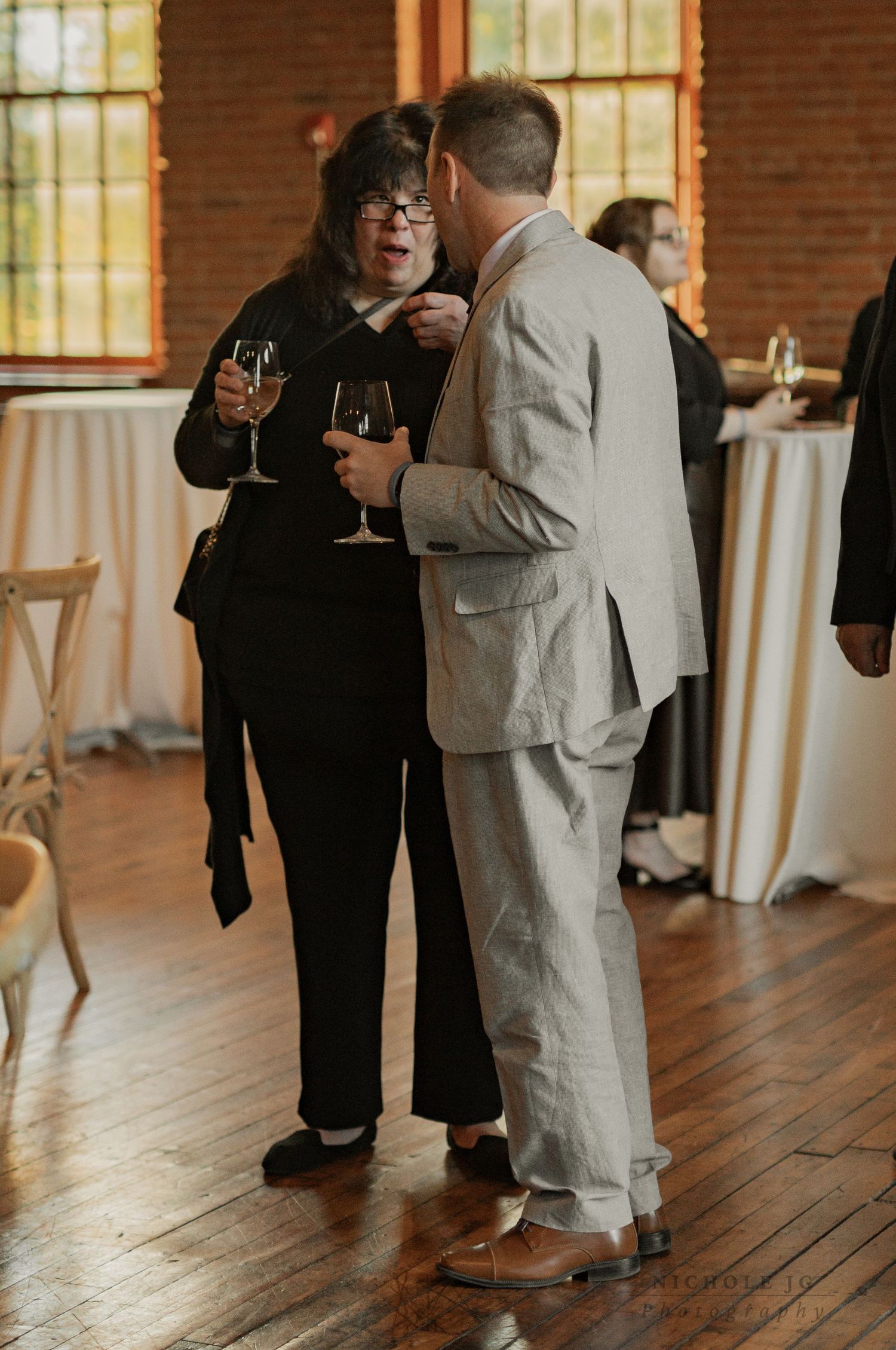 Two guests in formal attire share a conversation while holding wine glasses at an elegant indoor wedding reception.